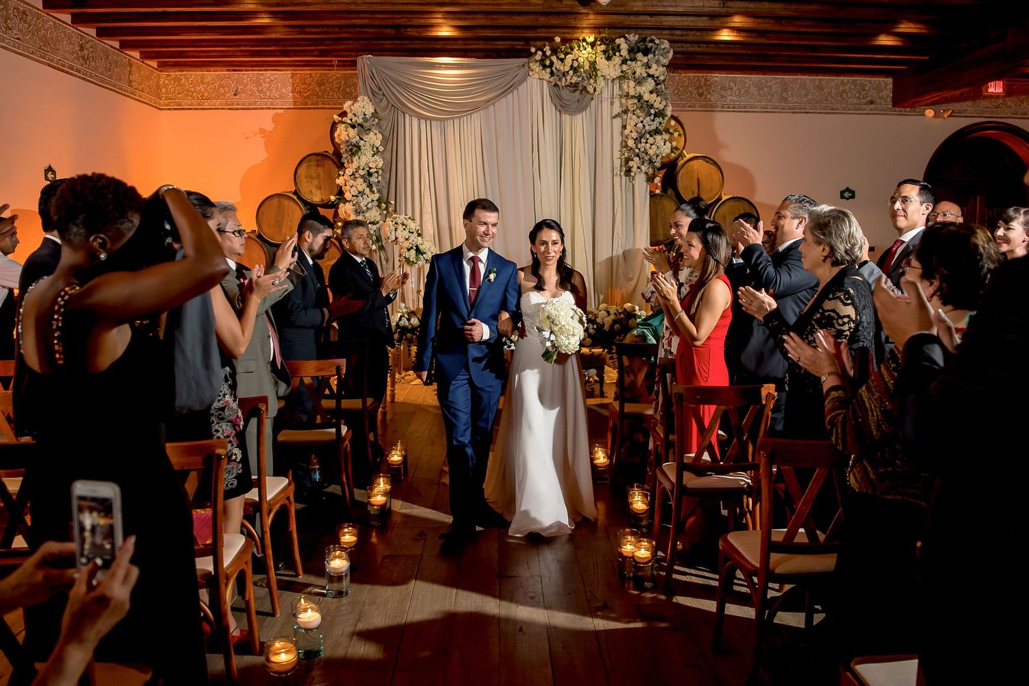 Bride and groom walk down the aisle together at an elegant indoor wedding ceremony with candlelit chairs.