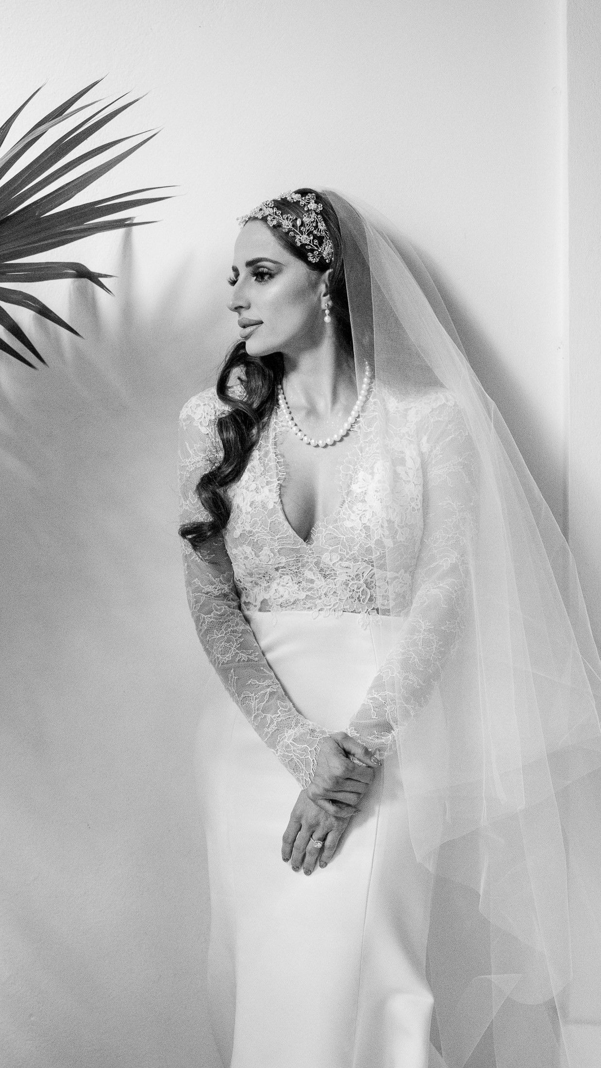 Black and white portrait of a bride in lace gown, pearl necklace, jeweled headpiece, and flowing veil.