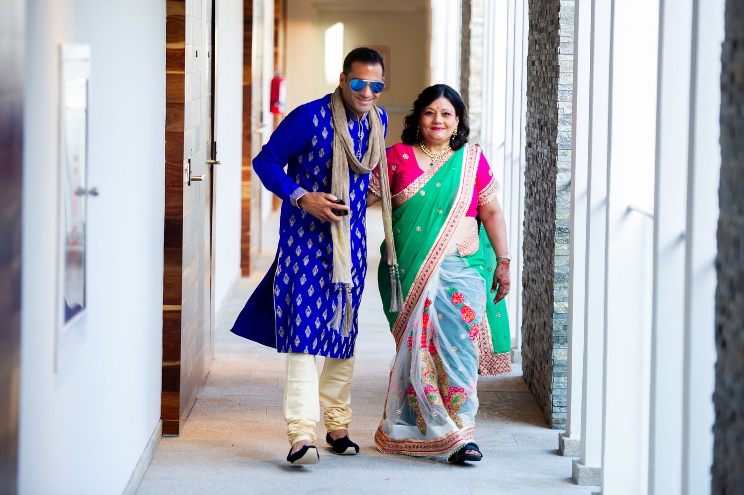 Couple in vibrant Indian attire, man in blue kurta and woman in colorful saree, walking elegantly in a hallway.