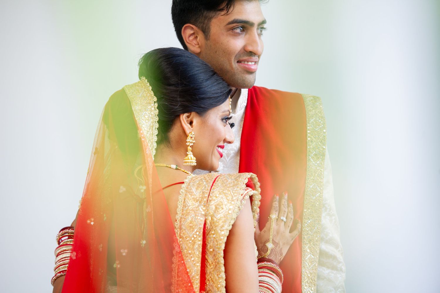 Indian bride and groom in traditional wedding attire, draped in red and gold fabric, sharing a romantic moment.