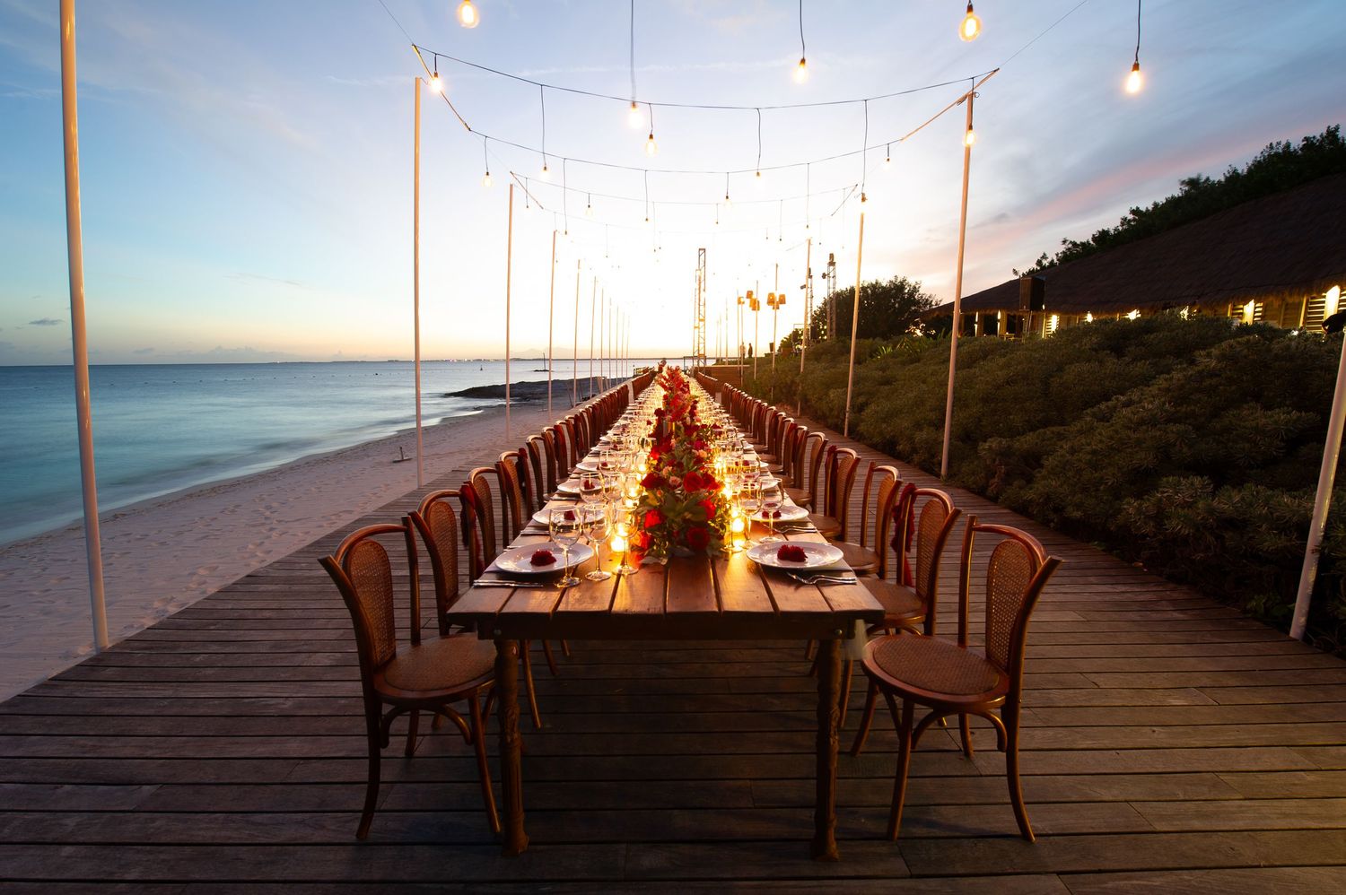 Elegant beachside dining table set for a sunset dinner party on a wooden deck with string lights overhead.