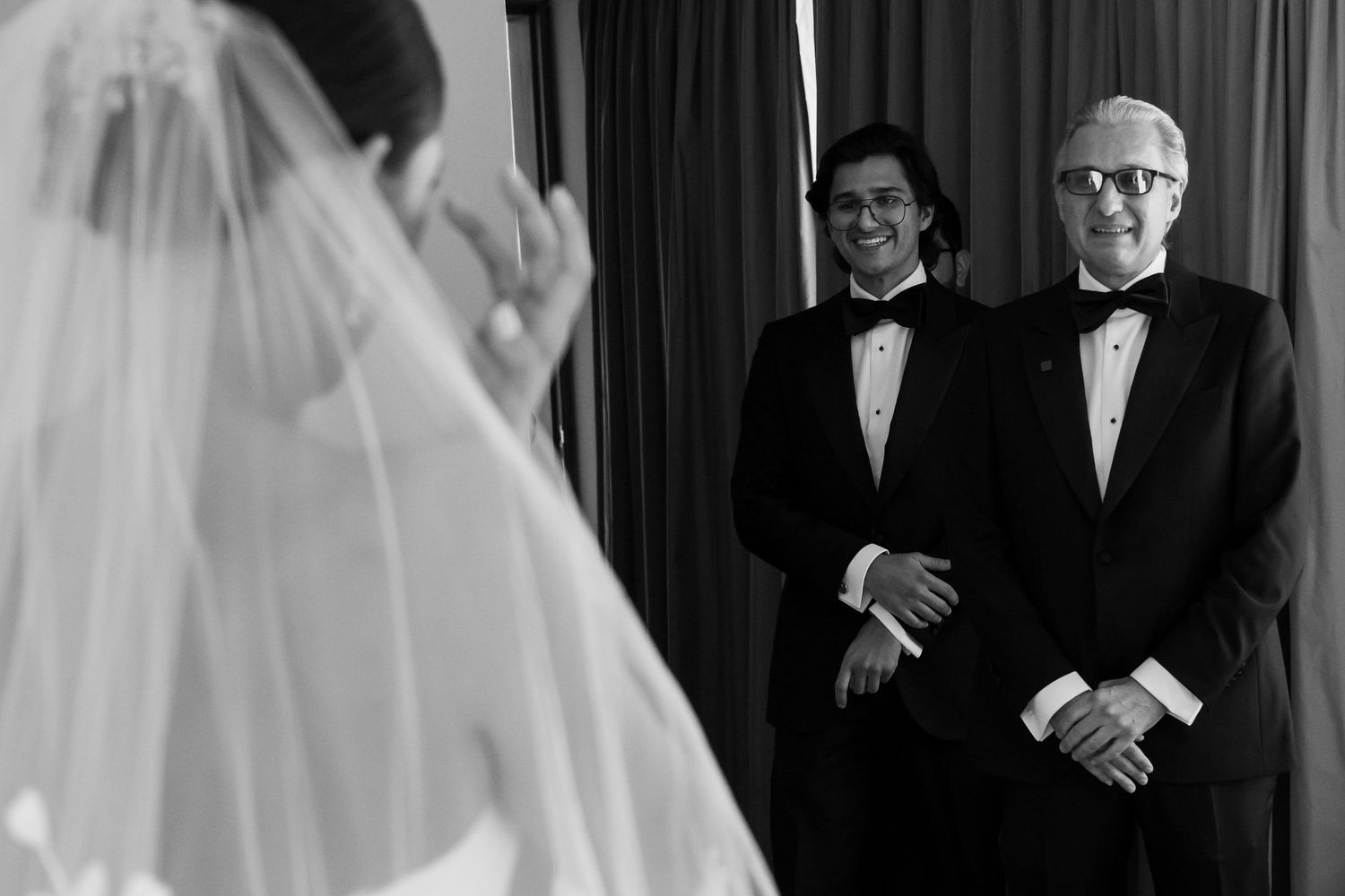 Black and white wedding ceremony photo of groom and officiant smiling as bride approaches in veil.