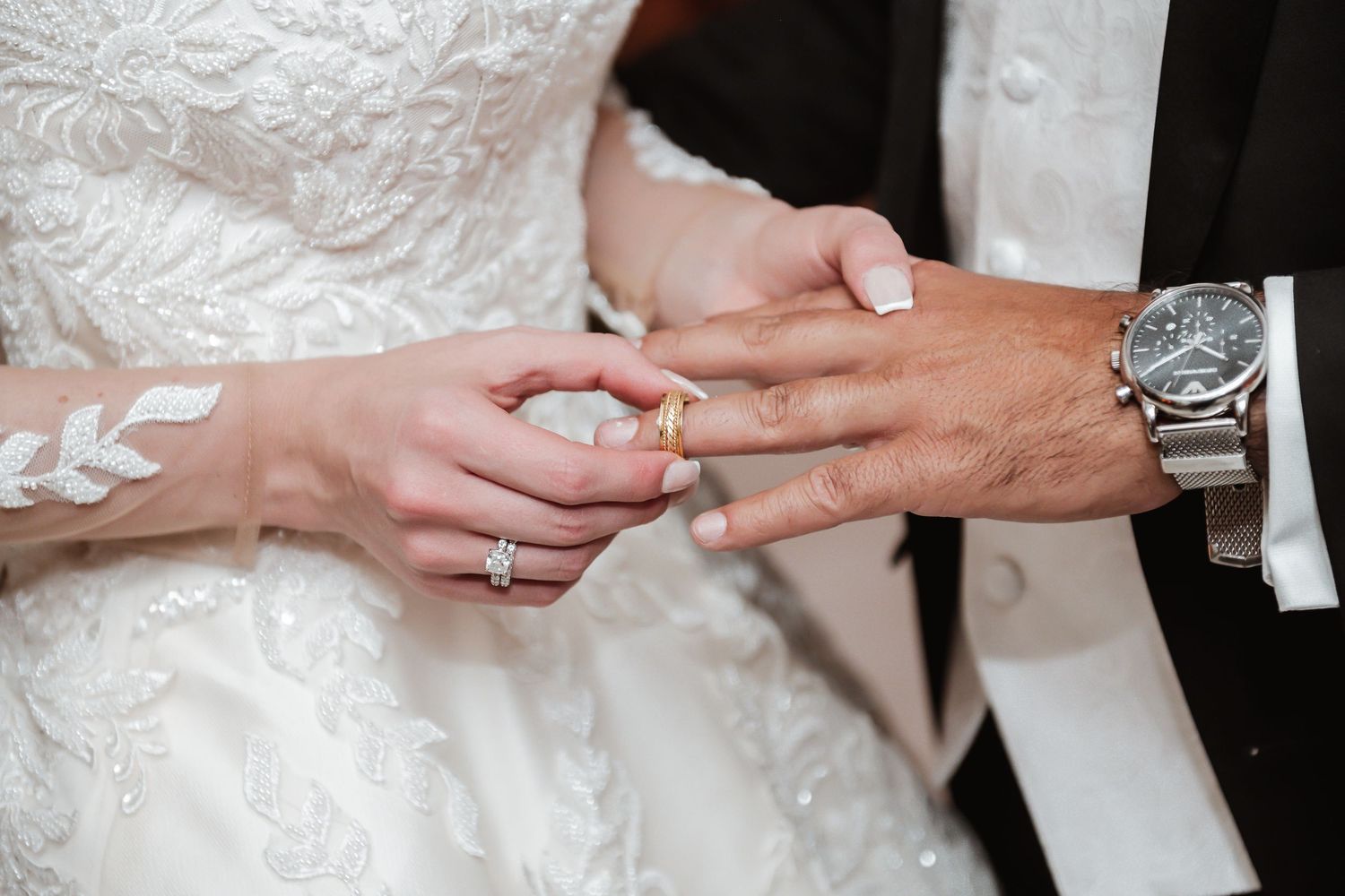 Close-up of wedding rings being exchanged with detailed lace dress and formal attire visible.
