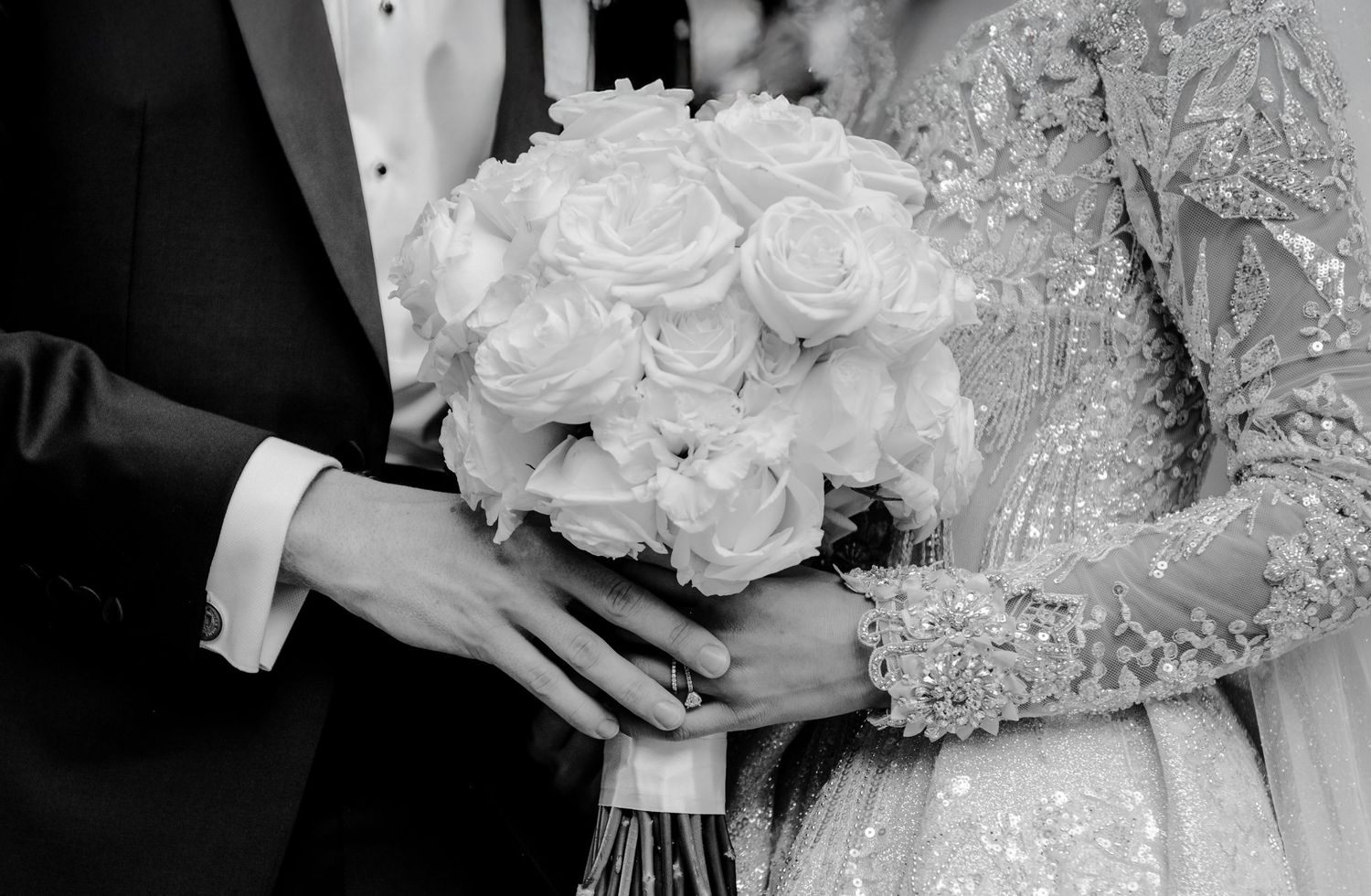 Black and white photo of bride and groom holding white rose wedding bouquet, close-up of hands in formal attire.