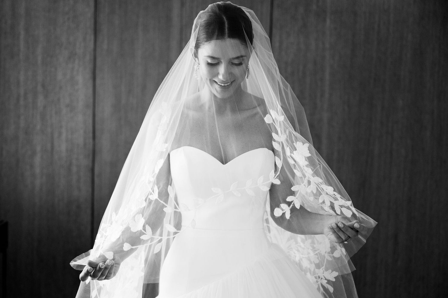 Black and white photo of a bride in a strapless gown and lace-trimmed veil, smiling softly while looking down.