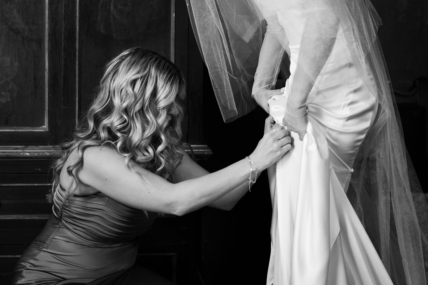 Bridesmaid kneeling to fasten wedding dress buttons on bride in elegant black and white photo.