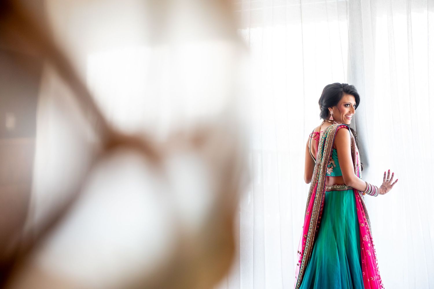Bride in teal and pink lehenga standing by window in soft natural light.