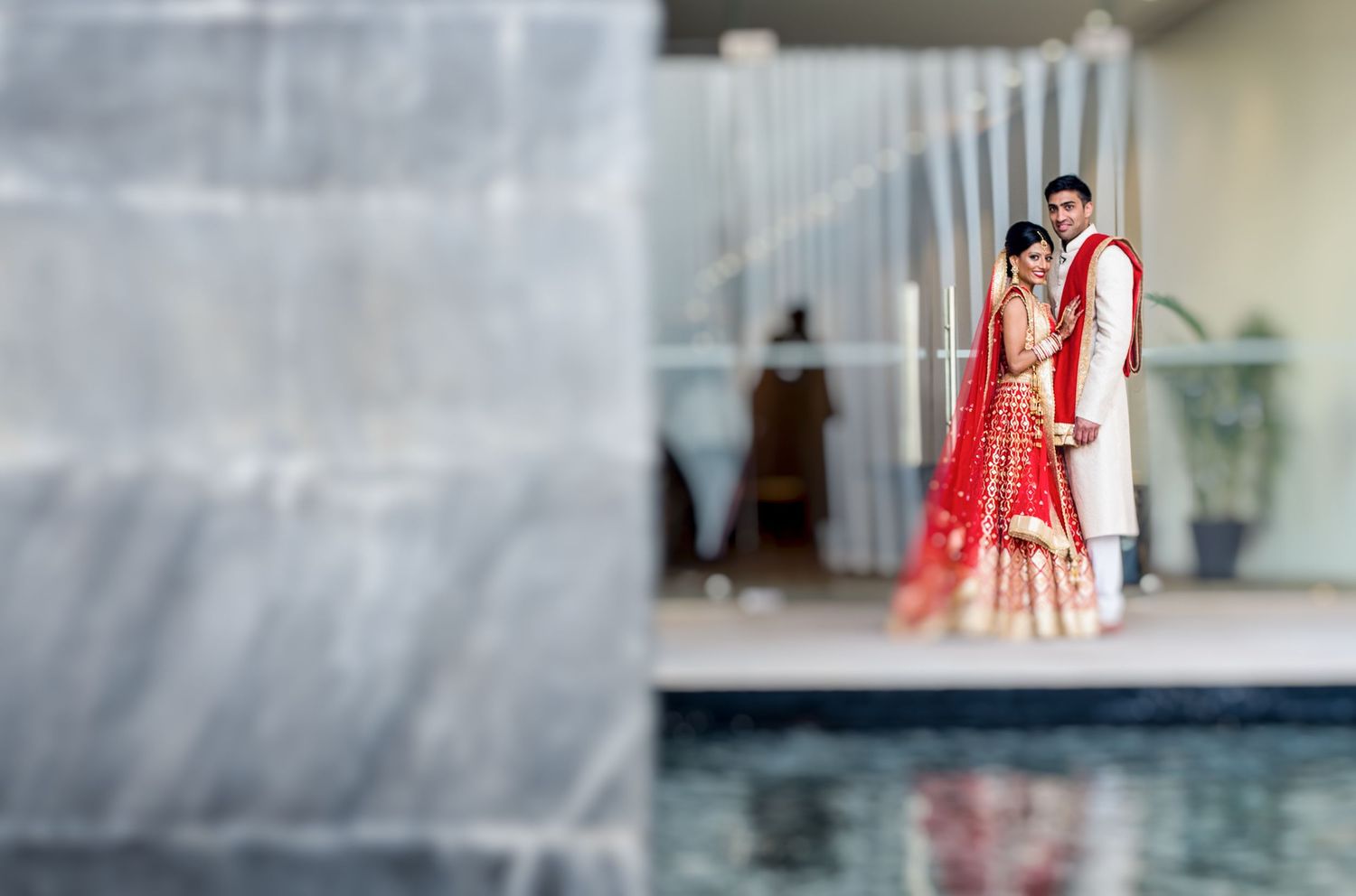 Indian bride in red lehenga and groom in white sherwani standing by a pool at their traditional wedding ceremony.