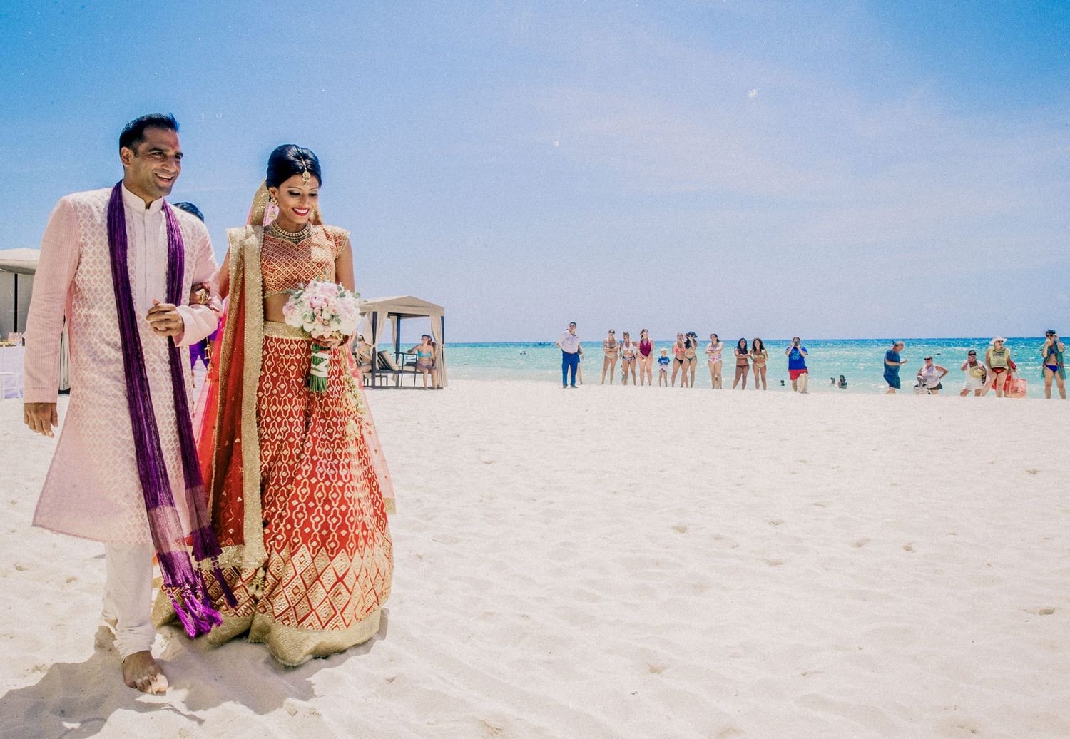 Indian couple in traditional wedding attire walking on a white sand beach with turquoise ocean backdrop.