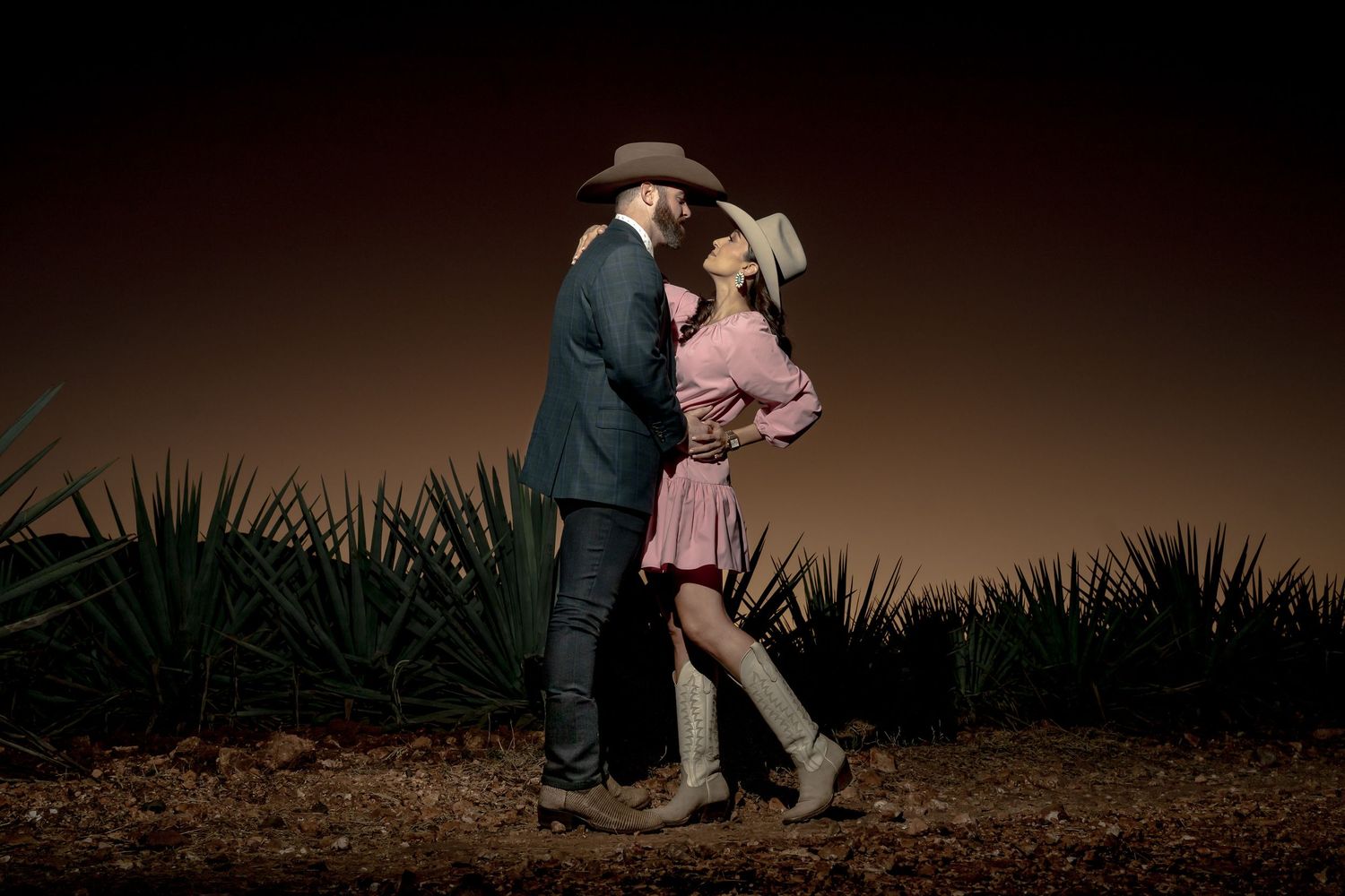 Couple in cowboy hats sharing a romantic moment at night among agave plants in a desert setting.