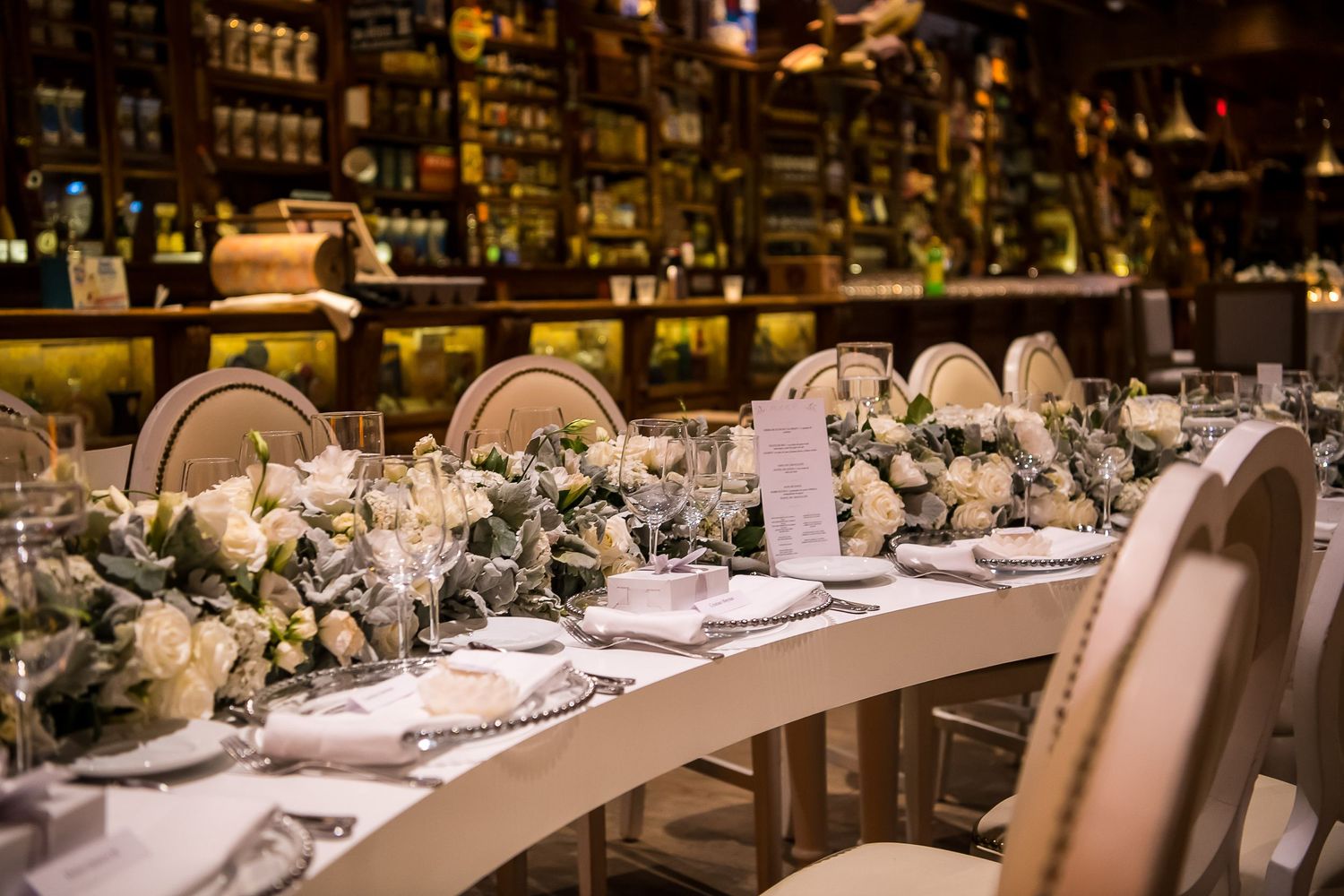 Elegant wedding reception table with white roses, greenery garland, fine china, and crystal glasses in a warmly lit restaurant.