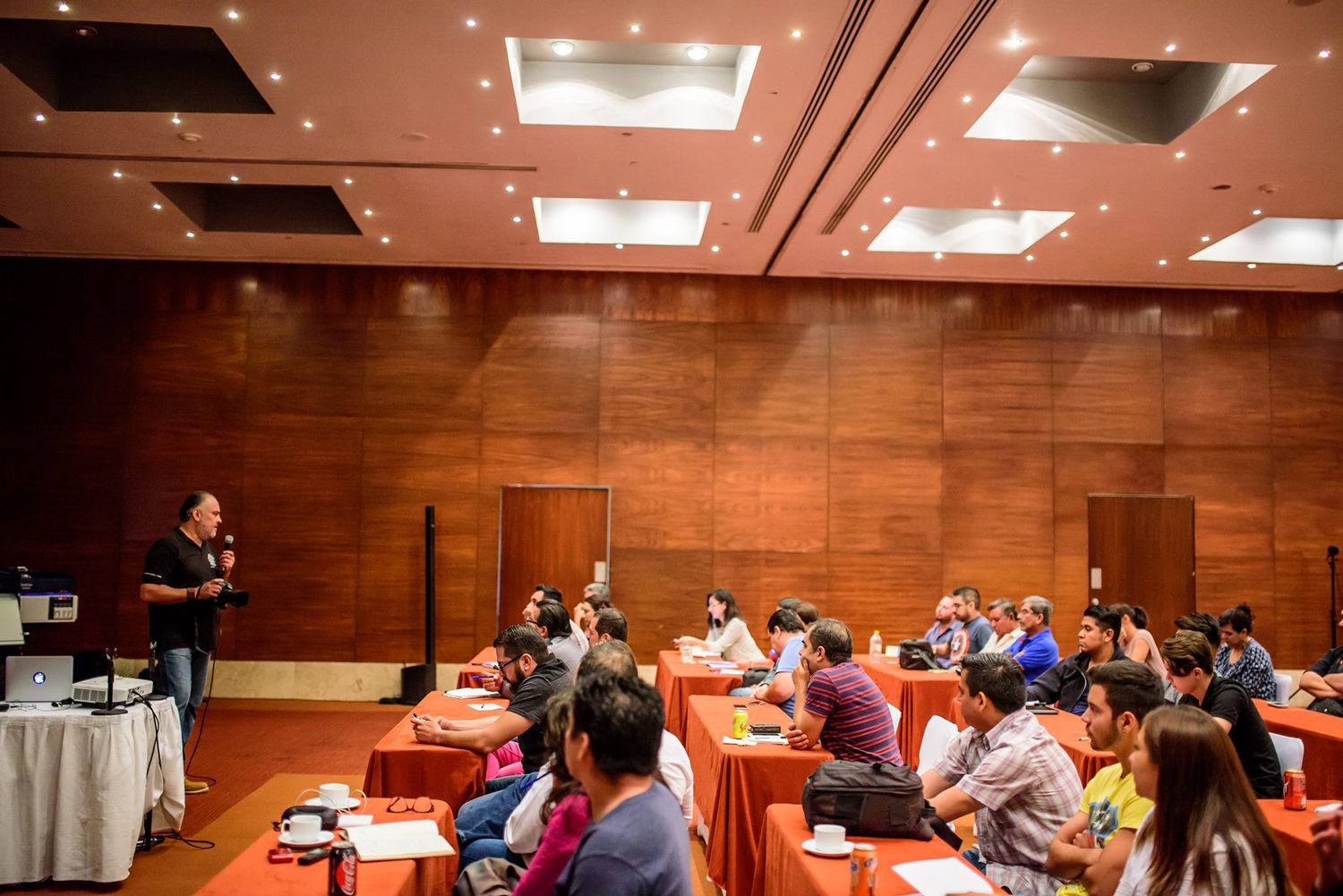Panoramic view of a conference hall with wooden walls and attendees seated at round tables.