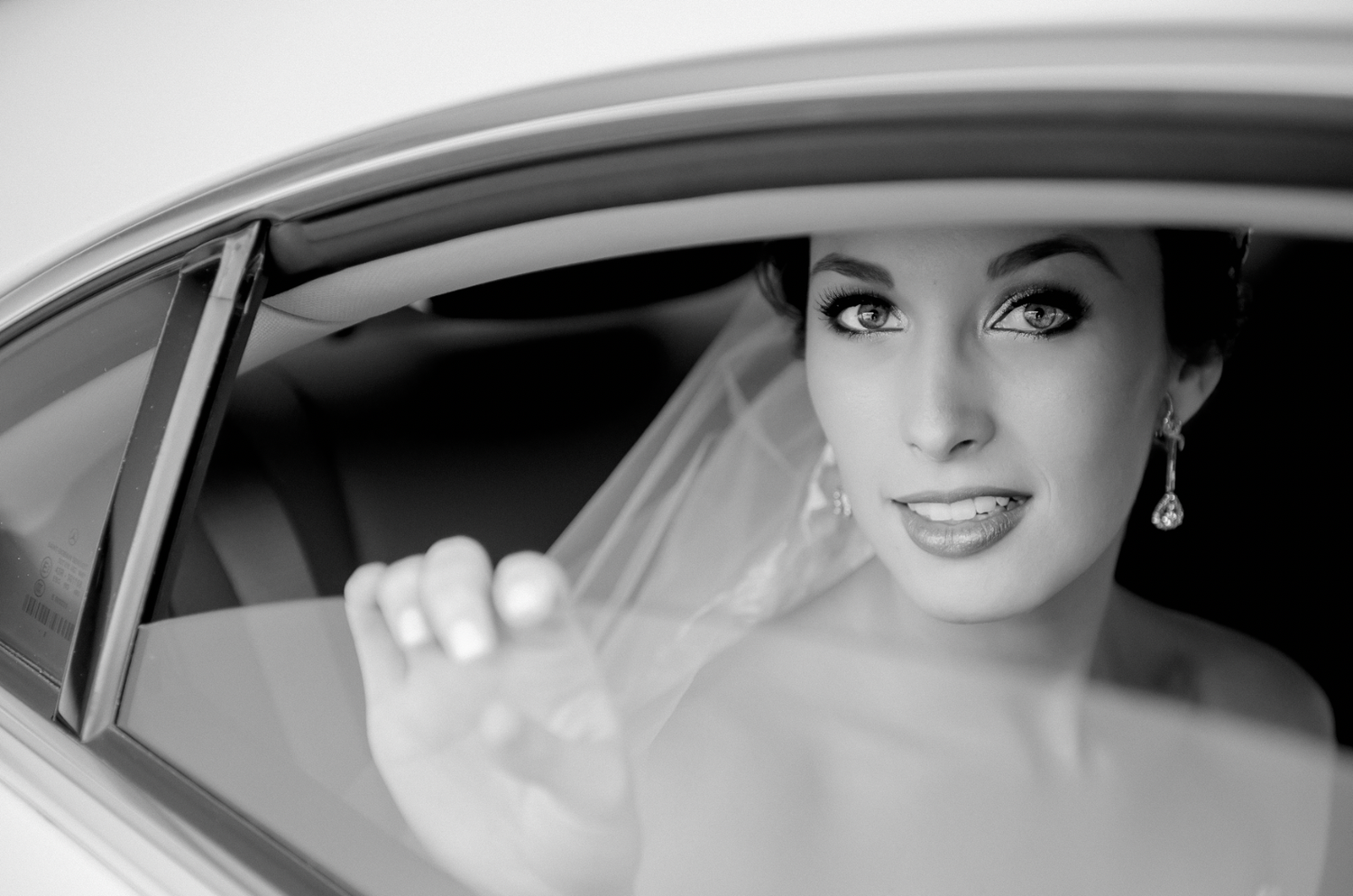 Dramatic black and white close-up shot through a car window showing bridal makeup and jewelry.