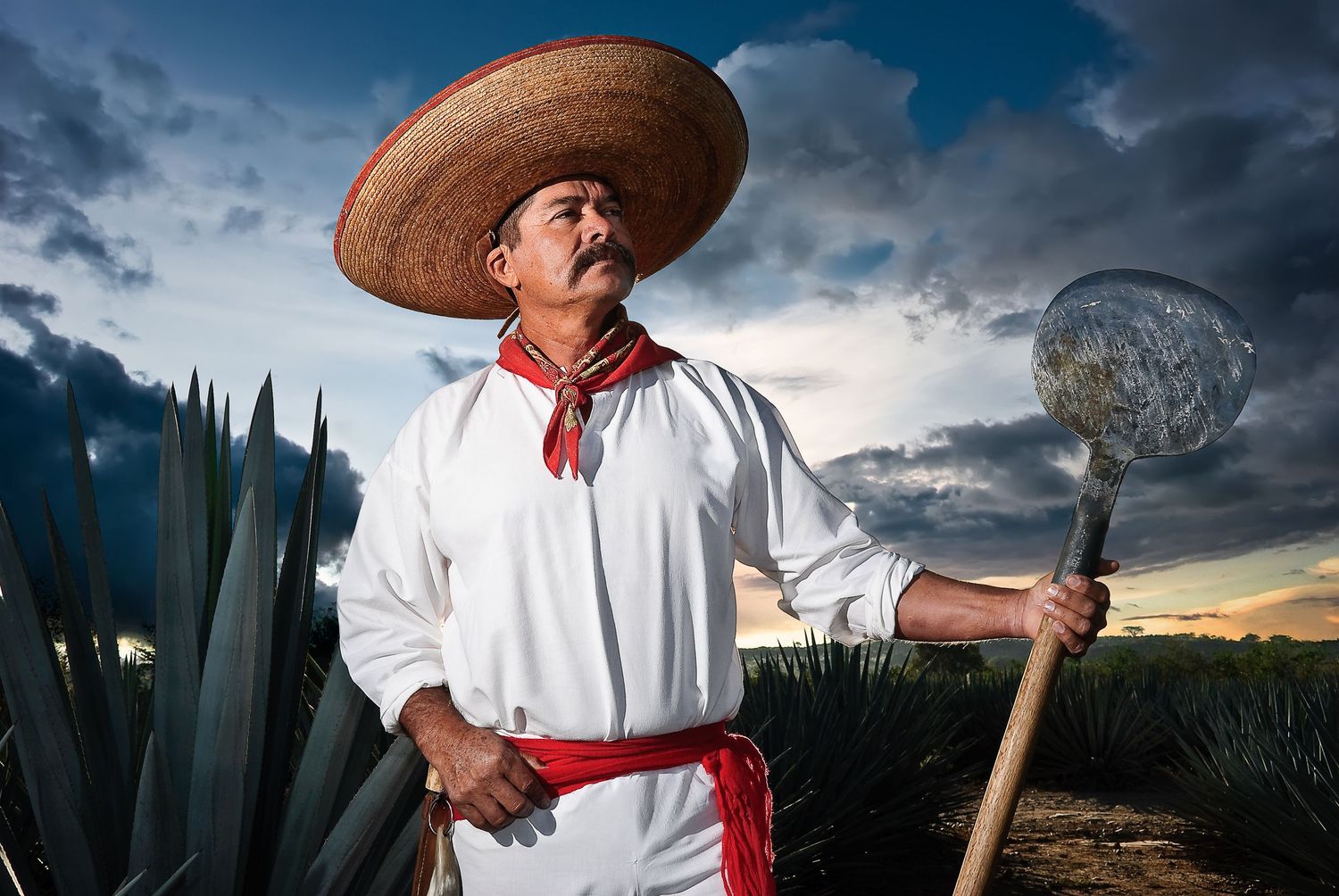 A traditional Mexican farmer in white clothing and sombrero poses with tools against blue sky.