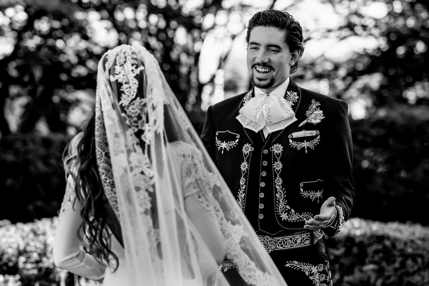 Black and white photo of a smiling groom in charro suit with bride in lace veil at a traditional Mexican wedding.