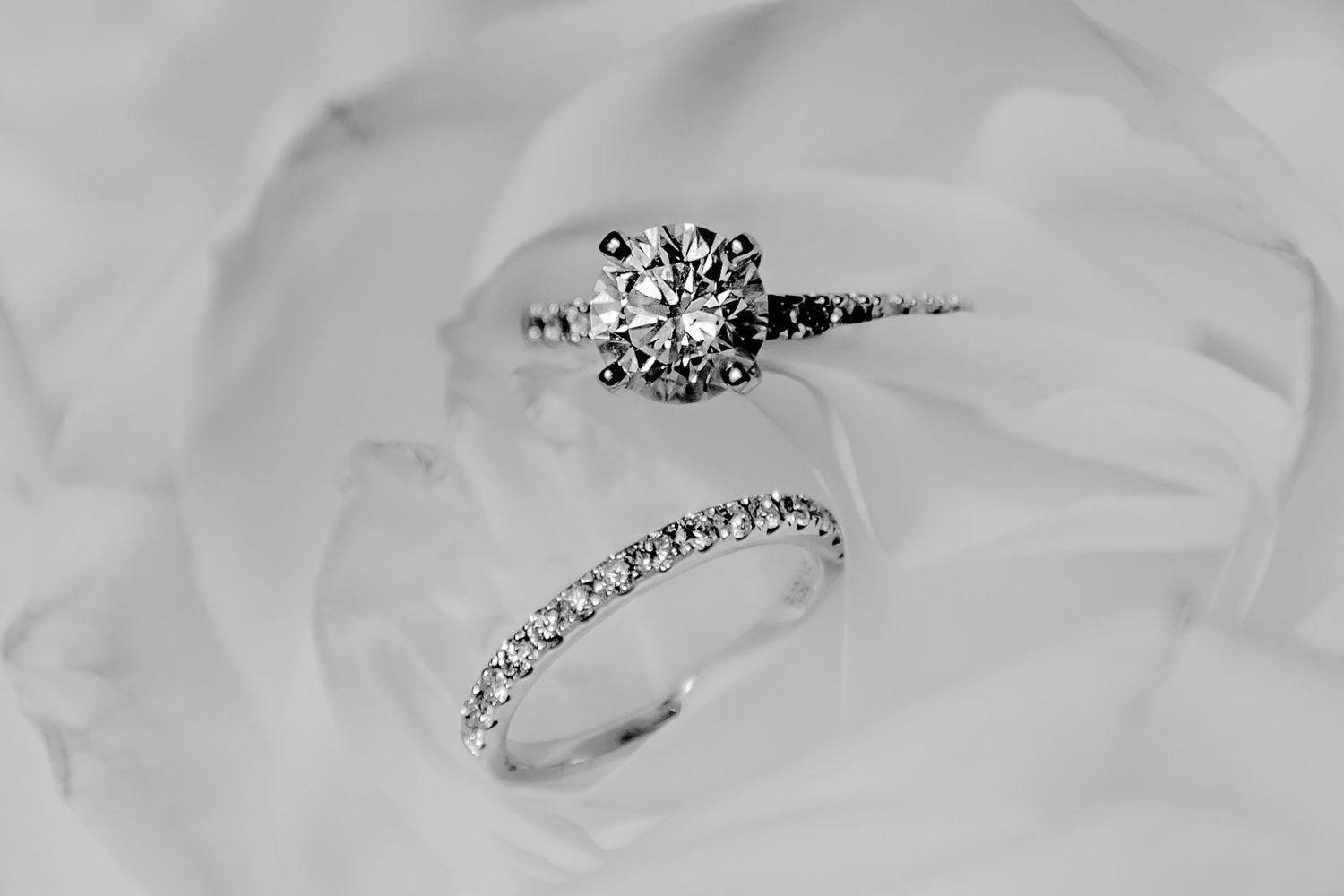 Elegant diamond engagement ring and wedding band set displayed on soft white fabric in black and white photography.