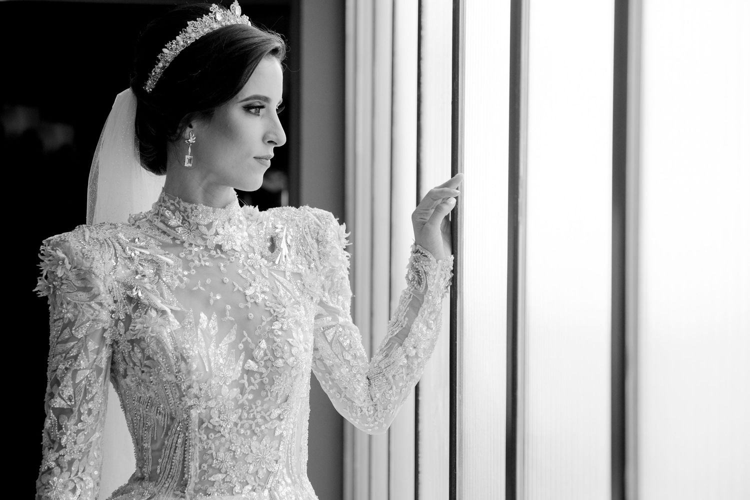 Elegant bride in a beaded long-sleeve wedding gown and tiara gazes out a window in a black and white portrait.