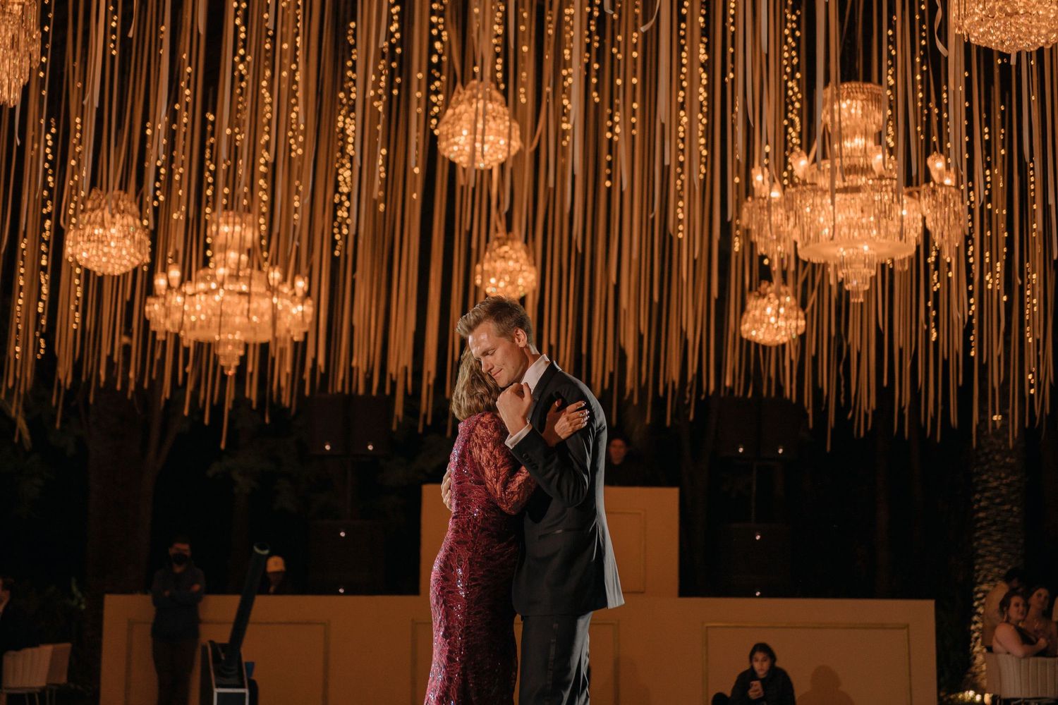 Couple sharing romantic first dance under crystal chandeliers and warm string lights at elegant wedding reception.