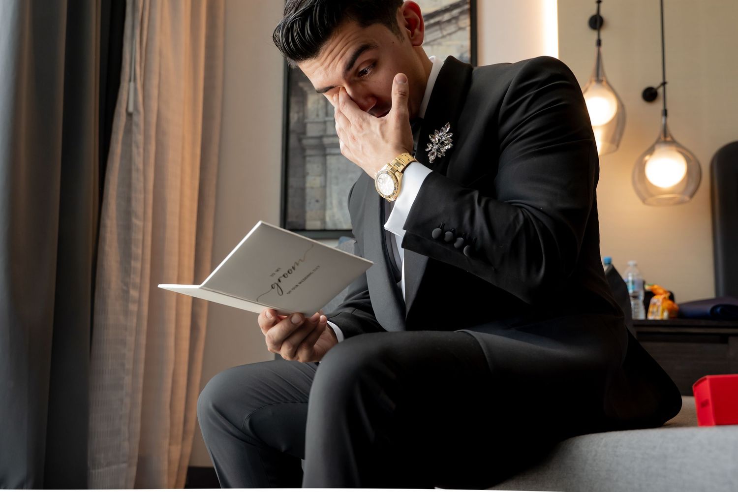Elegant man in black suit reading a card with a thoughtful expression, wearing a gold watch and boutonniere.