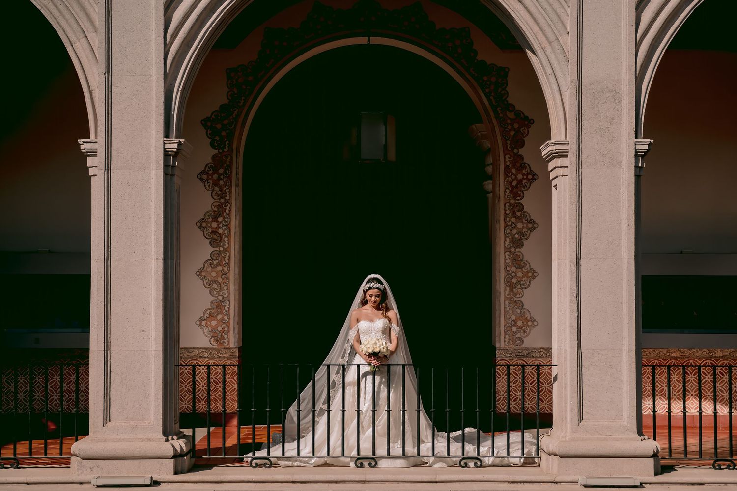 Bride in white gown and veil stands on ornate balcony between elegant arched columns at a historic venue.