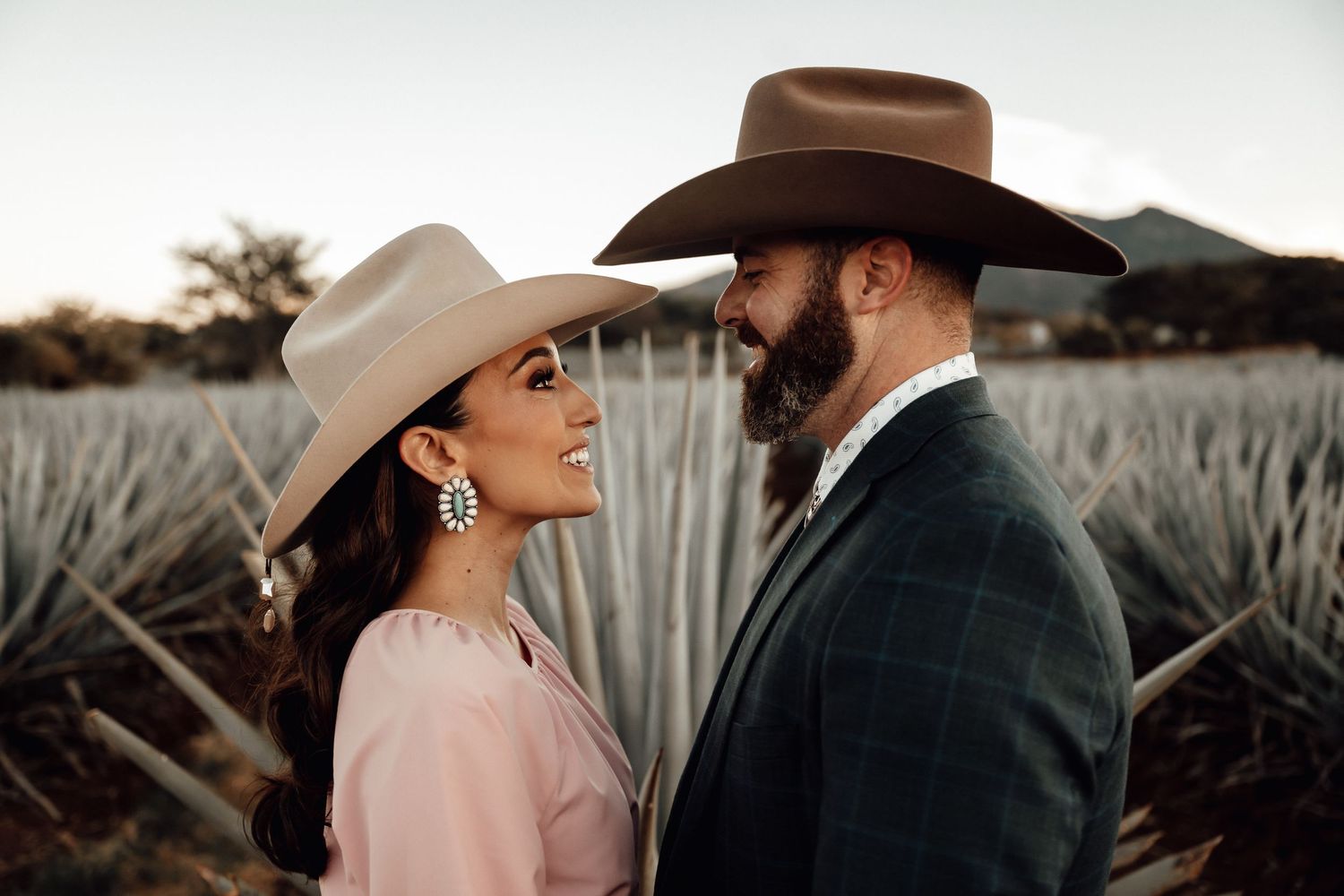 Couple in cowboy hats sharing a romantic moment in an agave field at sunset.