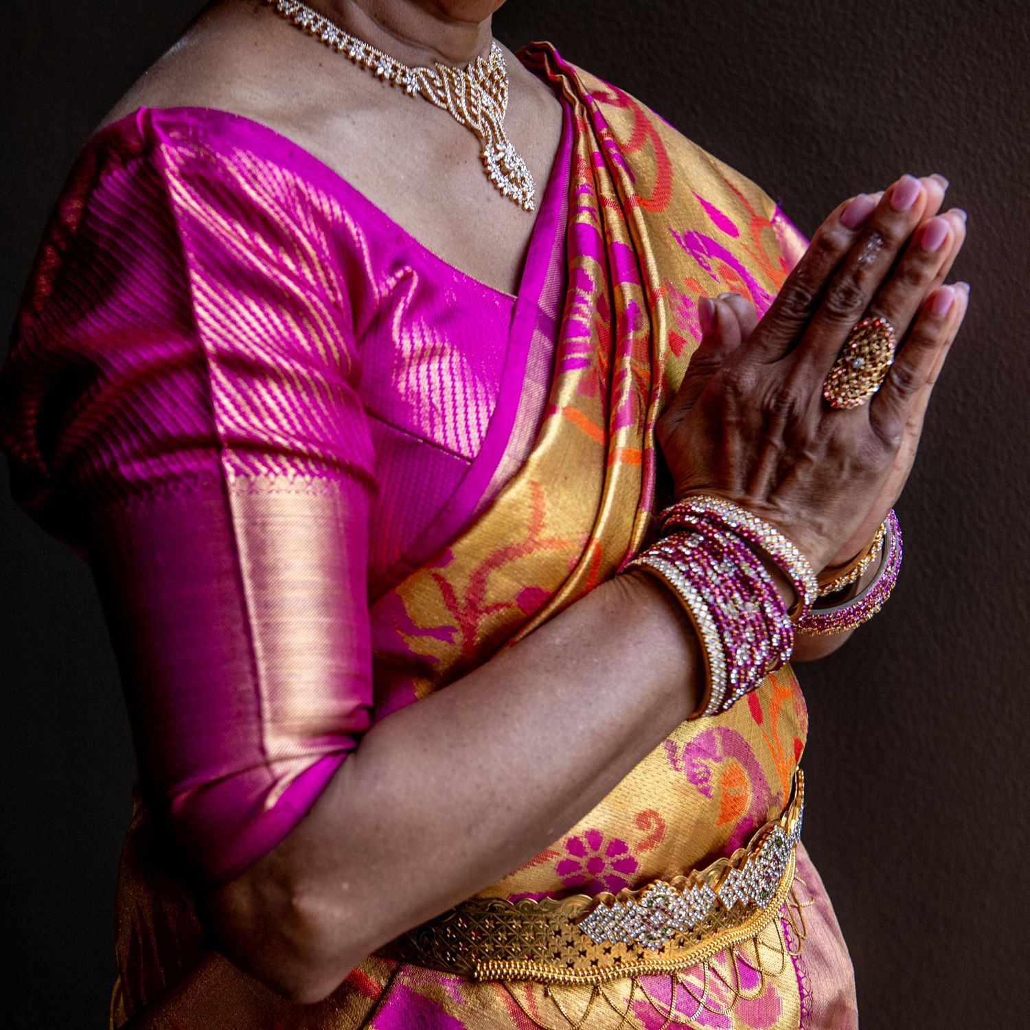 Woman in vibrant pink and gold silk saree performing namaste gesture, adorned with bangles and diamond jewelry.