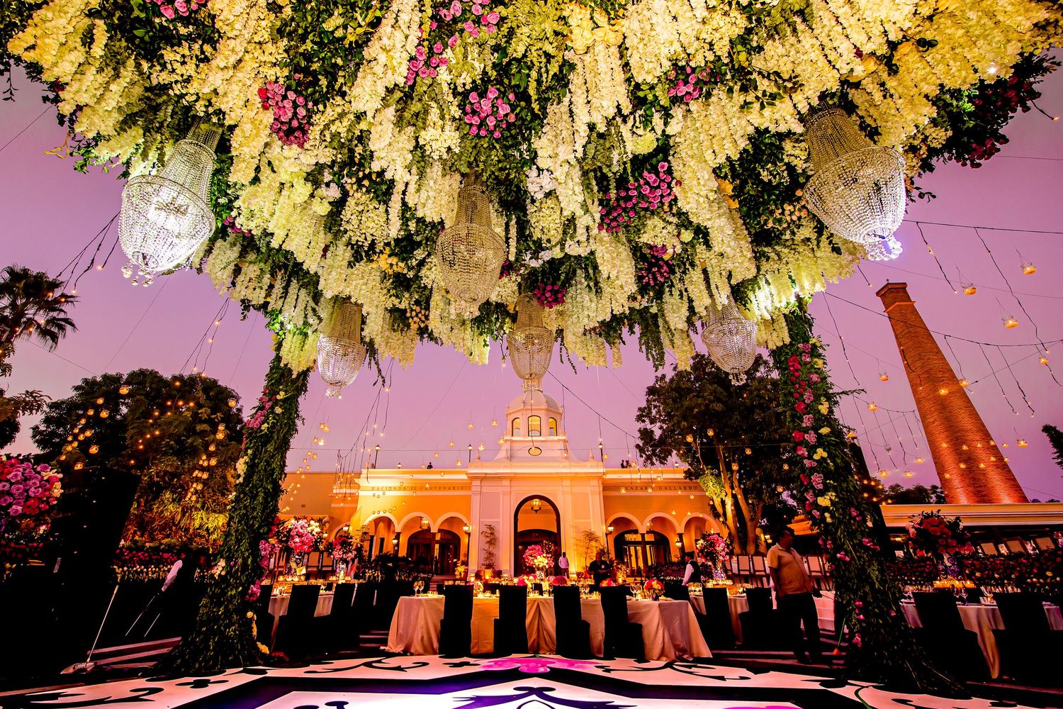 Yellow floral arrangements and chandeliers create a magical ceiling display at an outdoor evening event.