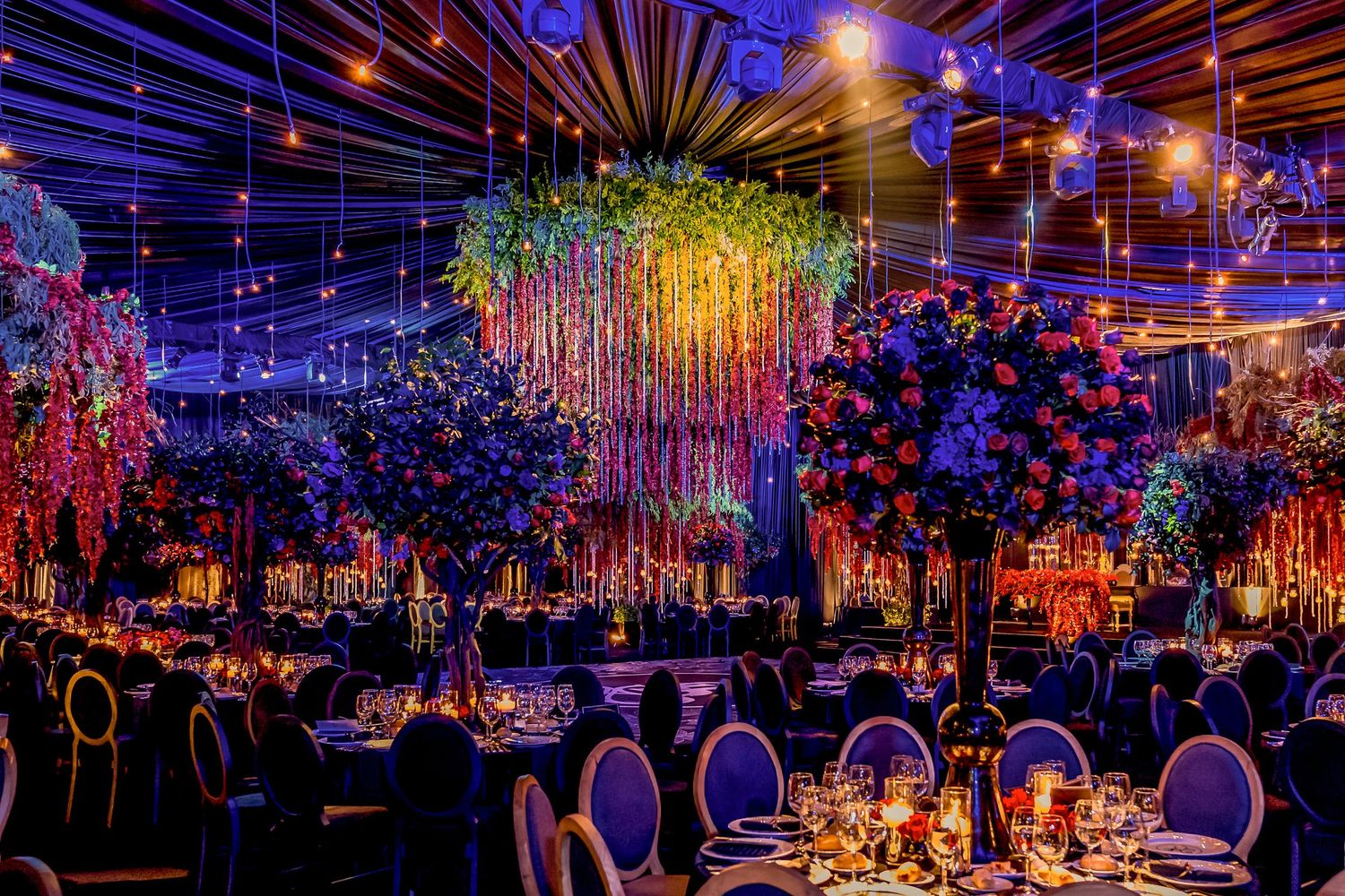 Enchanting wedding reception space with colorful hanging floral installations and dramatic lighting effects.
