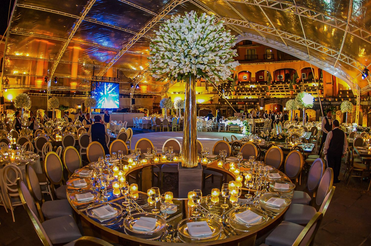 Elegant wedding reception hall with tall white floral centerpieces and round tables illuminated by warm candlelight.