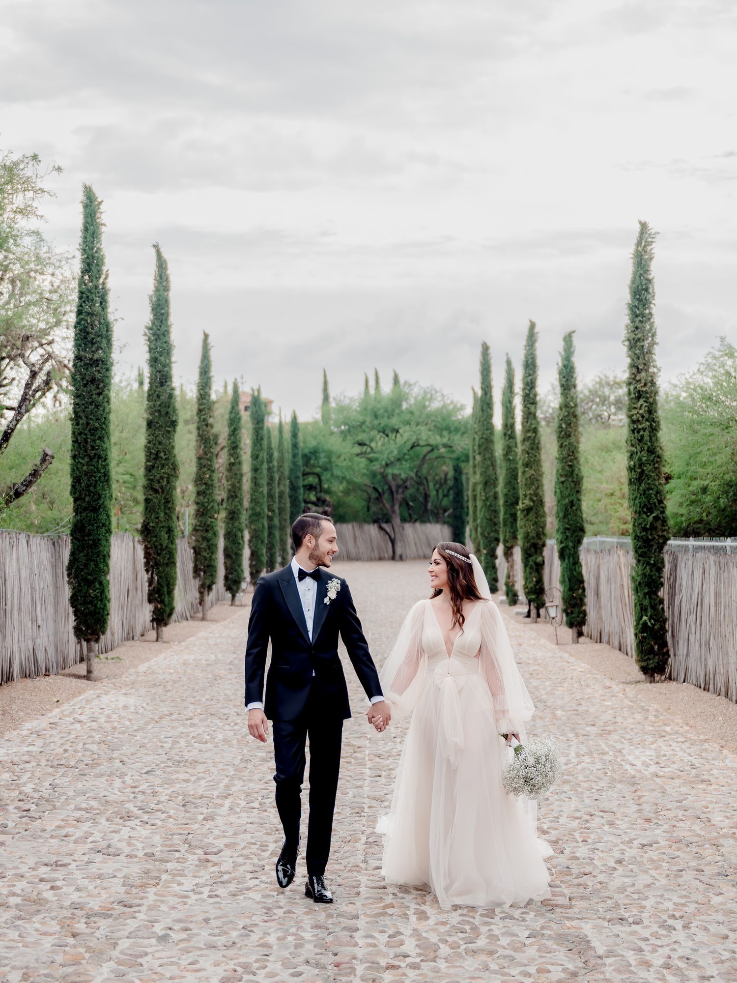 A romantic couple strolls along a cypress tree-lined path in an elegant wedding setting on a cloudy day.