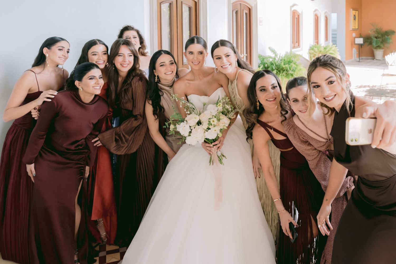 Bride in white gown holds bouquet surrounded by bridesmaids in brown and burgundy dresses posing for a group selfie.