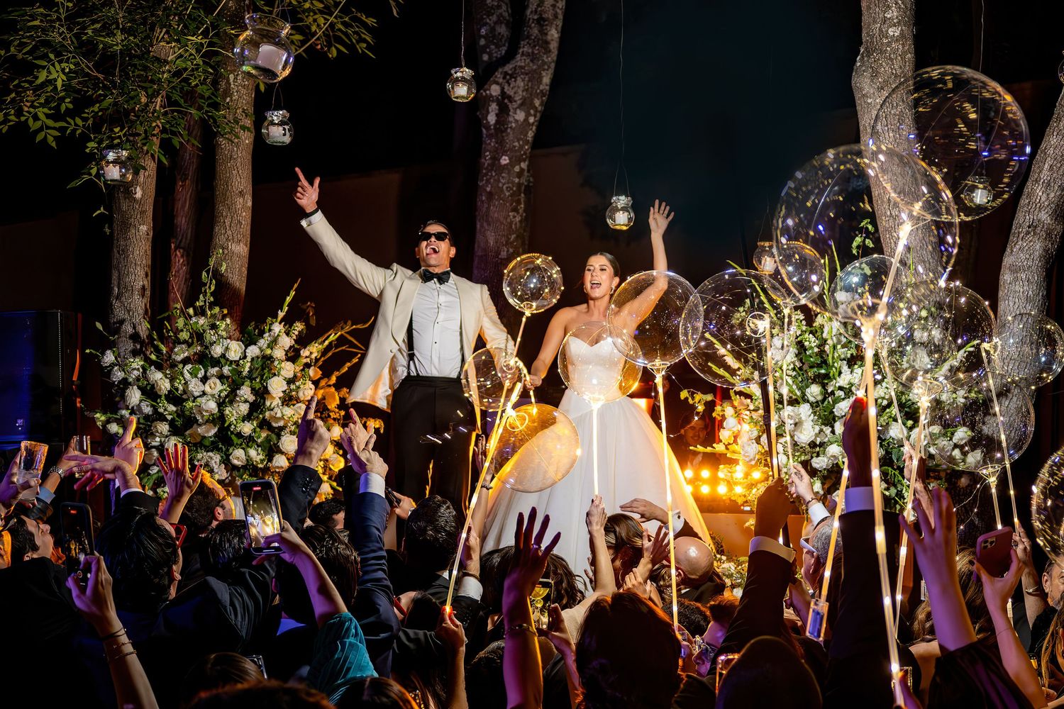 Bride and groom celebrate on elevated platform at night wedding reception with glowing balloons and cheering guests.