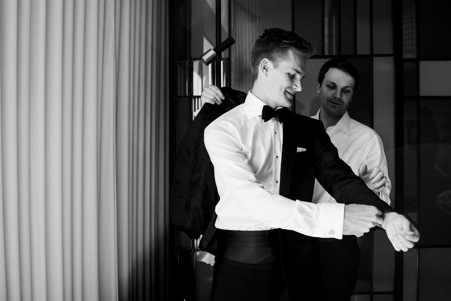 Groom adjusting cufflinks with groomsman assistance in black and white wedding preparation photo.