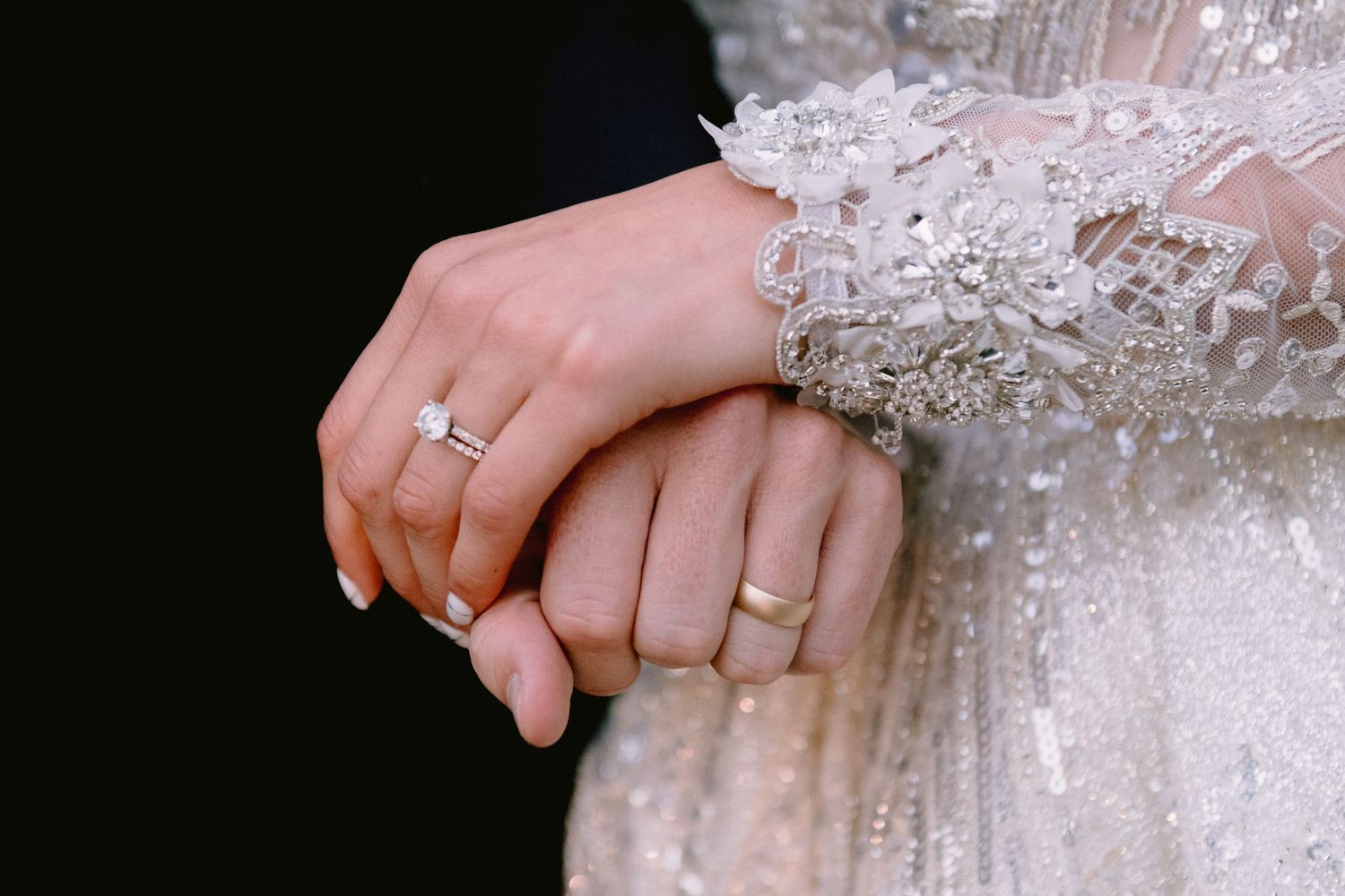 Close-up of bride and groom hands showing diamond engagement ring and gold wedding band on wedding day.