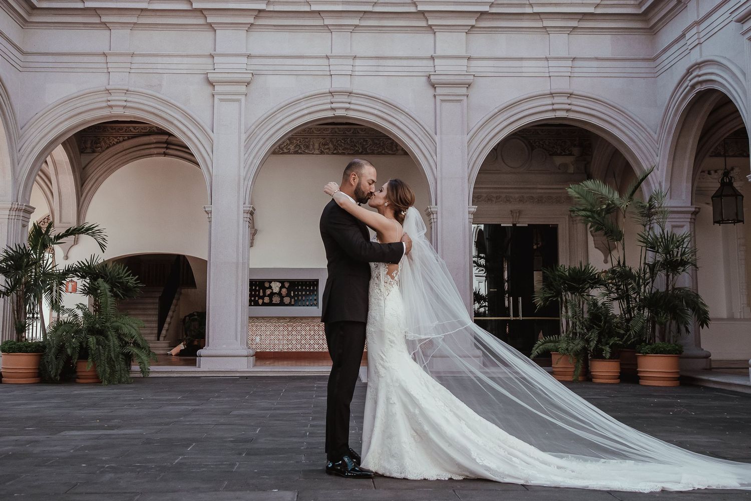 Bride and groom share romantic embrace in elegant arched courtyard, cathedral veil flowing behind white wedding gown.