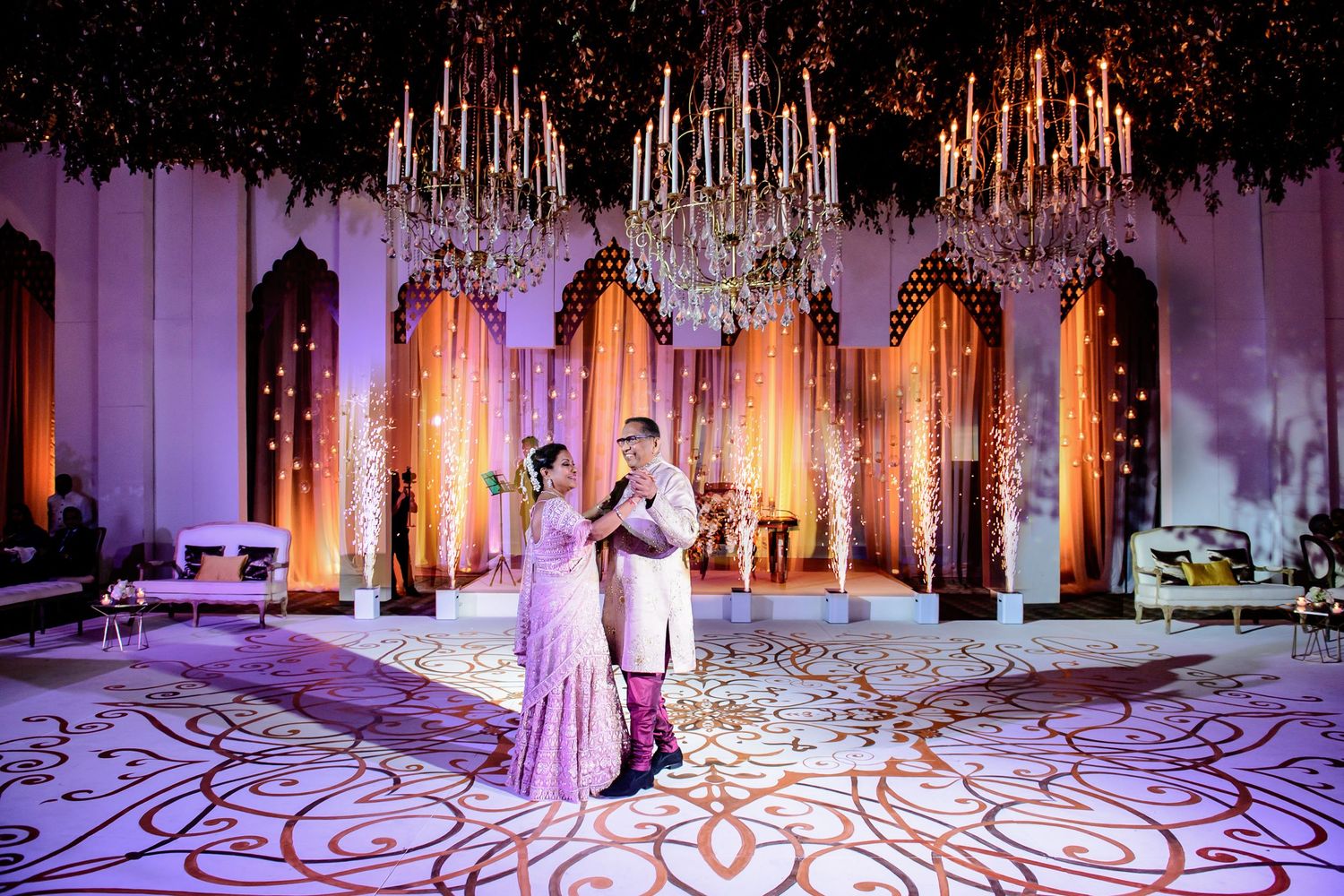 Couple in traditional Indian attire dancing at an elegant wedding venue with chandeliers and floral decor.