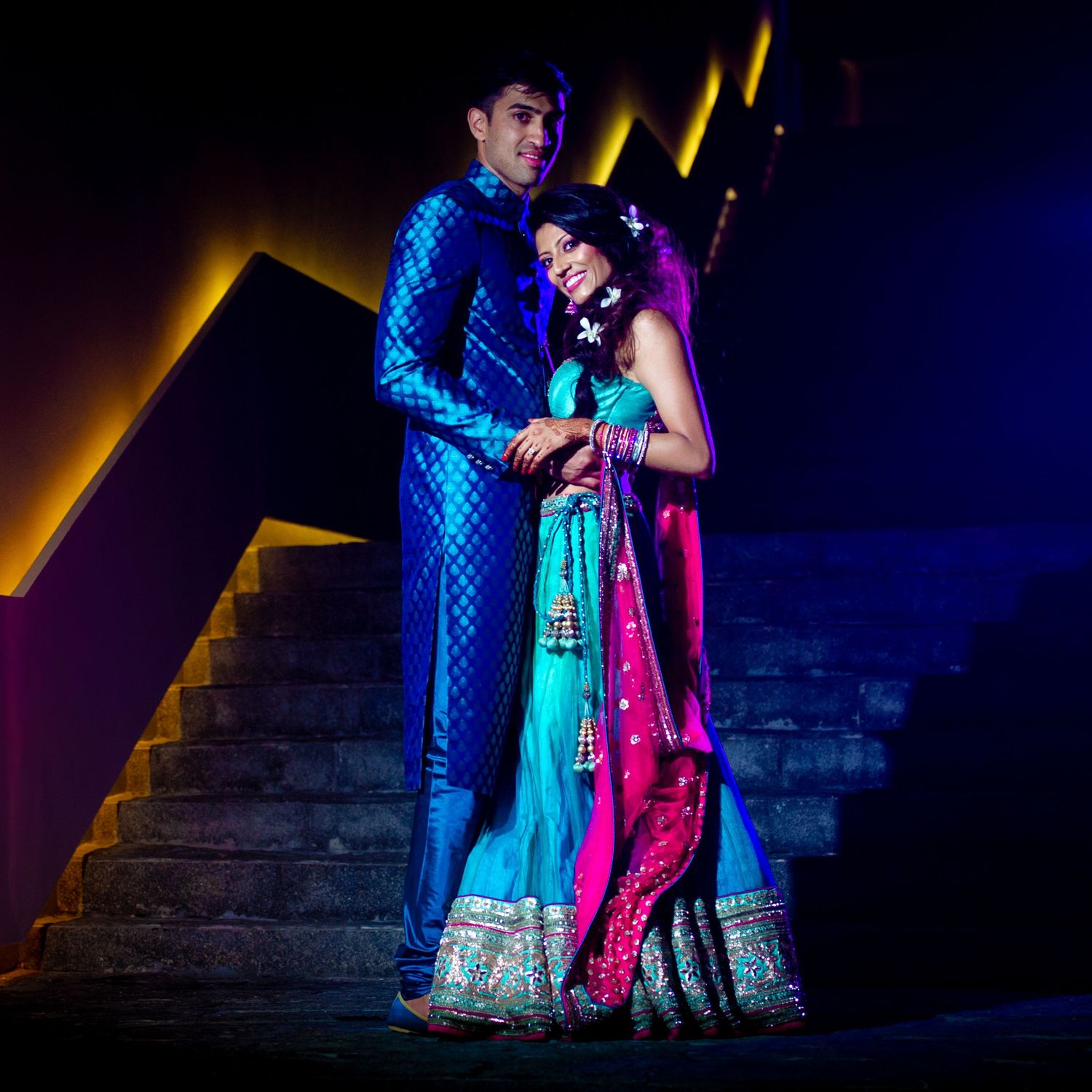 Indian couple in vibrant blue and pink traditional wedding attire posing elegantly on decorative stairs with colorful lighting.