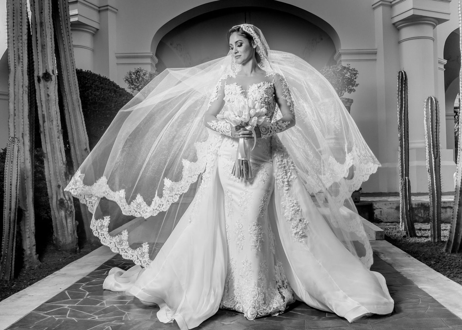 Elegant bride in lace mermaid wedding gown with long sleeves and flowing veil, posing outdoors in black and white photo.