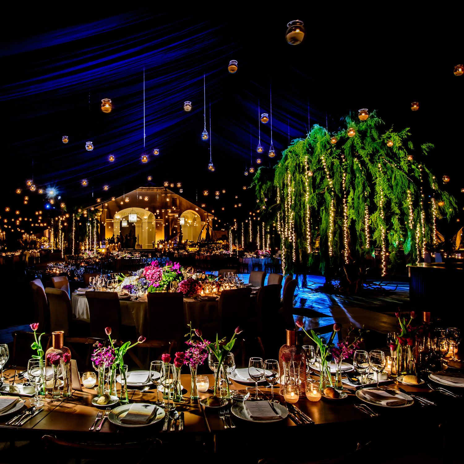 Elegant wedding reception venue with dramatic blue lighting and hanging floral arrangements illuminated by warm candlelight.