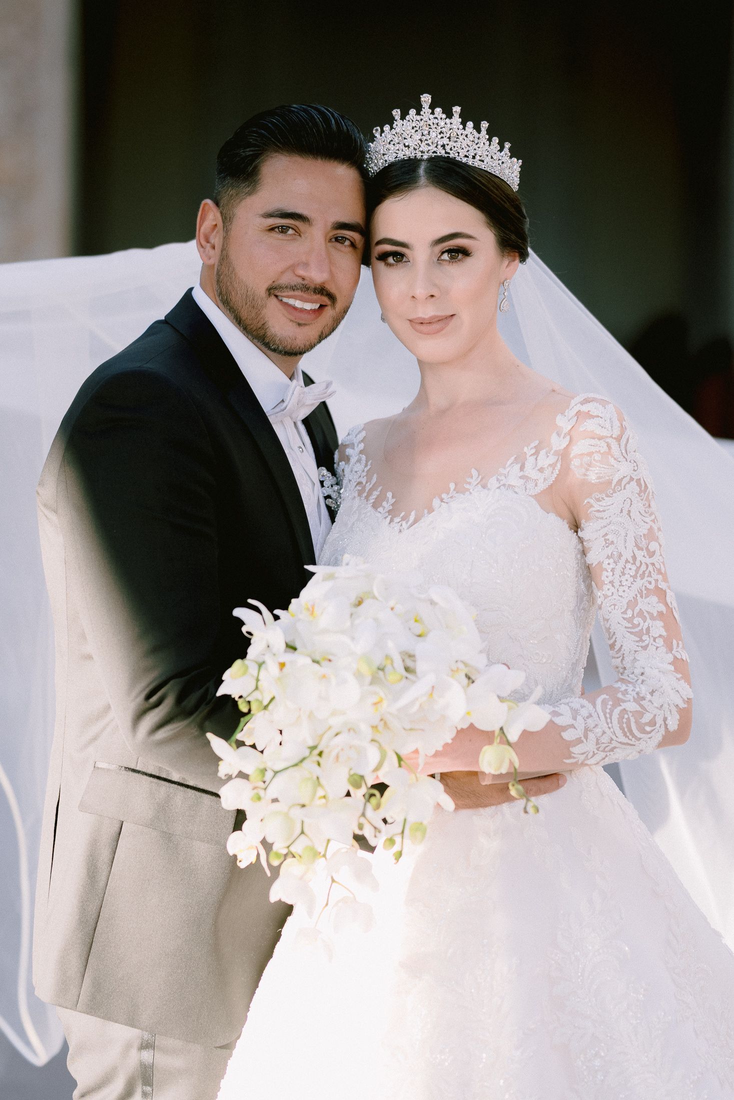 Elegant wedding portrait featuring a bride in a lace gown with tiara and groom in light-colored suit.