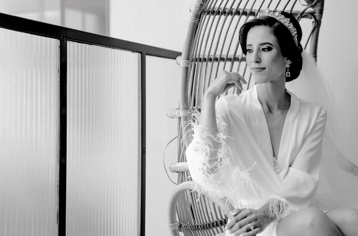 Elegant bride in white feathered robe and tiara sits in wicker chair, black and white portrait.
