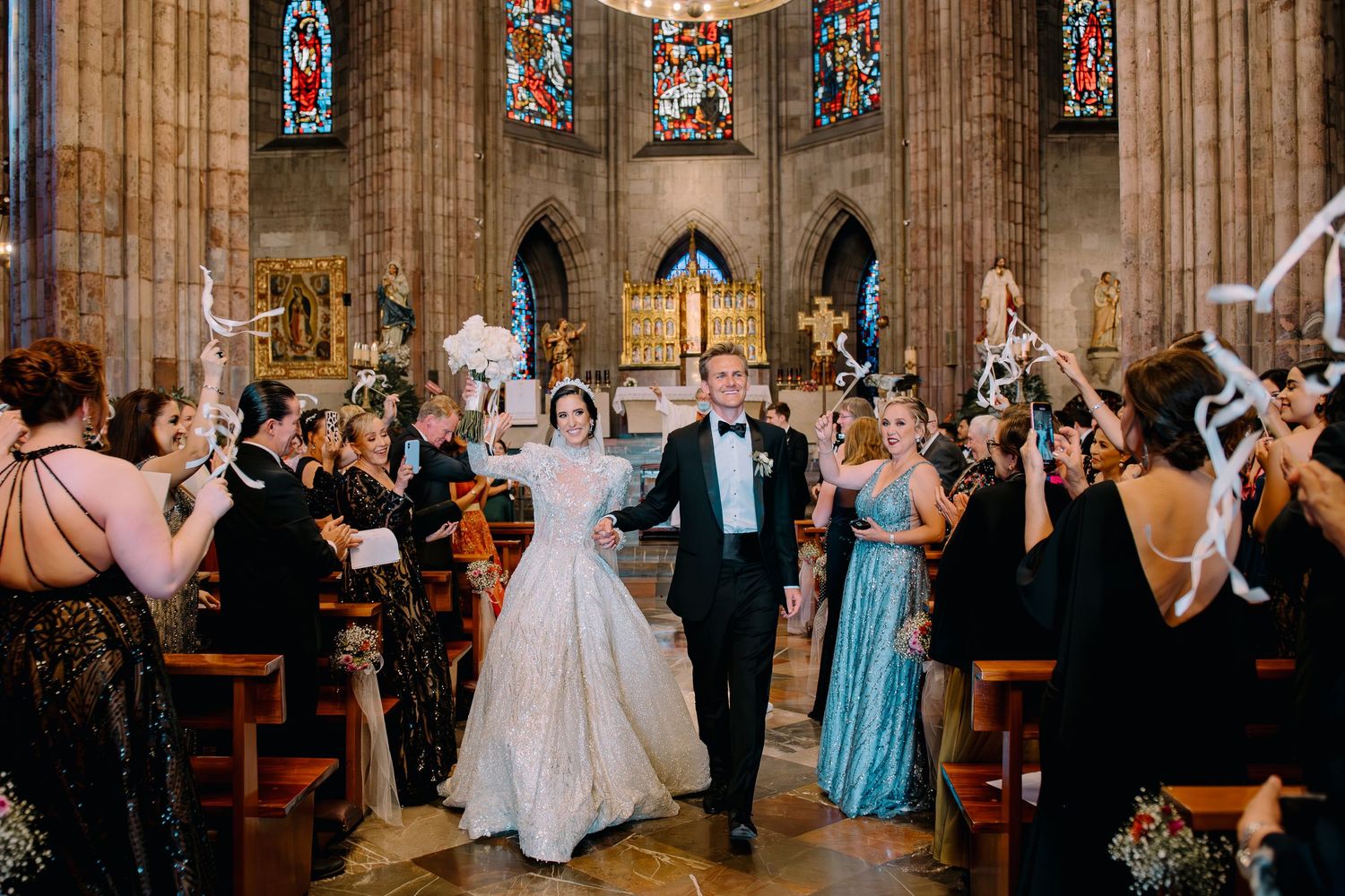 Bride and groom walk down cathedral aisle surrounded by cheering guests in elegant attire during wedding recessional.