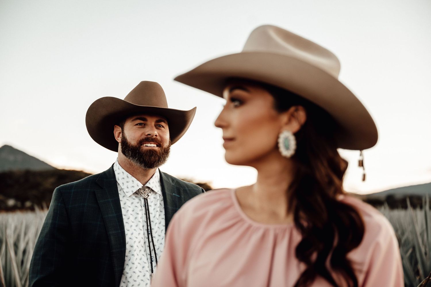 Couple in Western cowboy hats, man in dark suit with bolo tie, woman in pink blouse, posing outdoors at sunset.