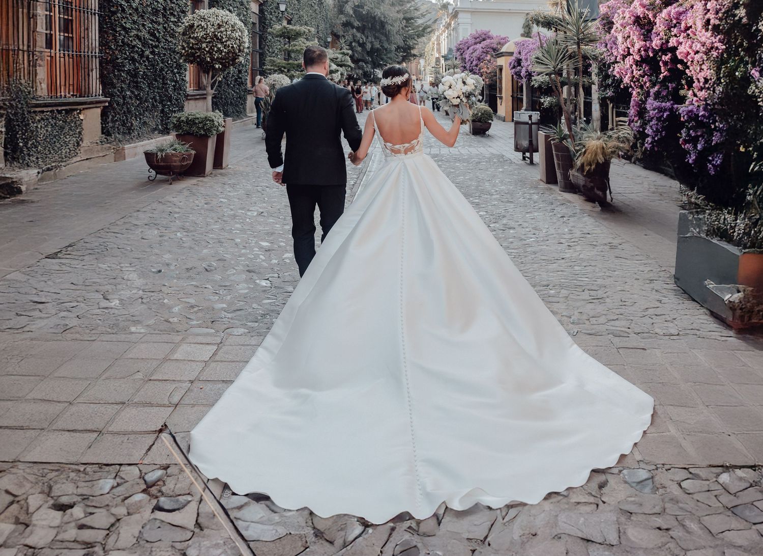 Bride and groom walking hand in hand on cobblestone path, elegant white ball gown with long train, floral backdrop.