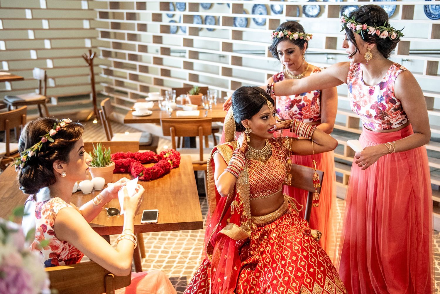 Indian bride in red lehenga gets ready with bridesmaids in pink floral outfits and flower crowns.