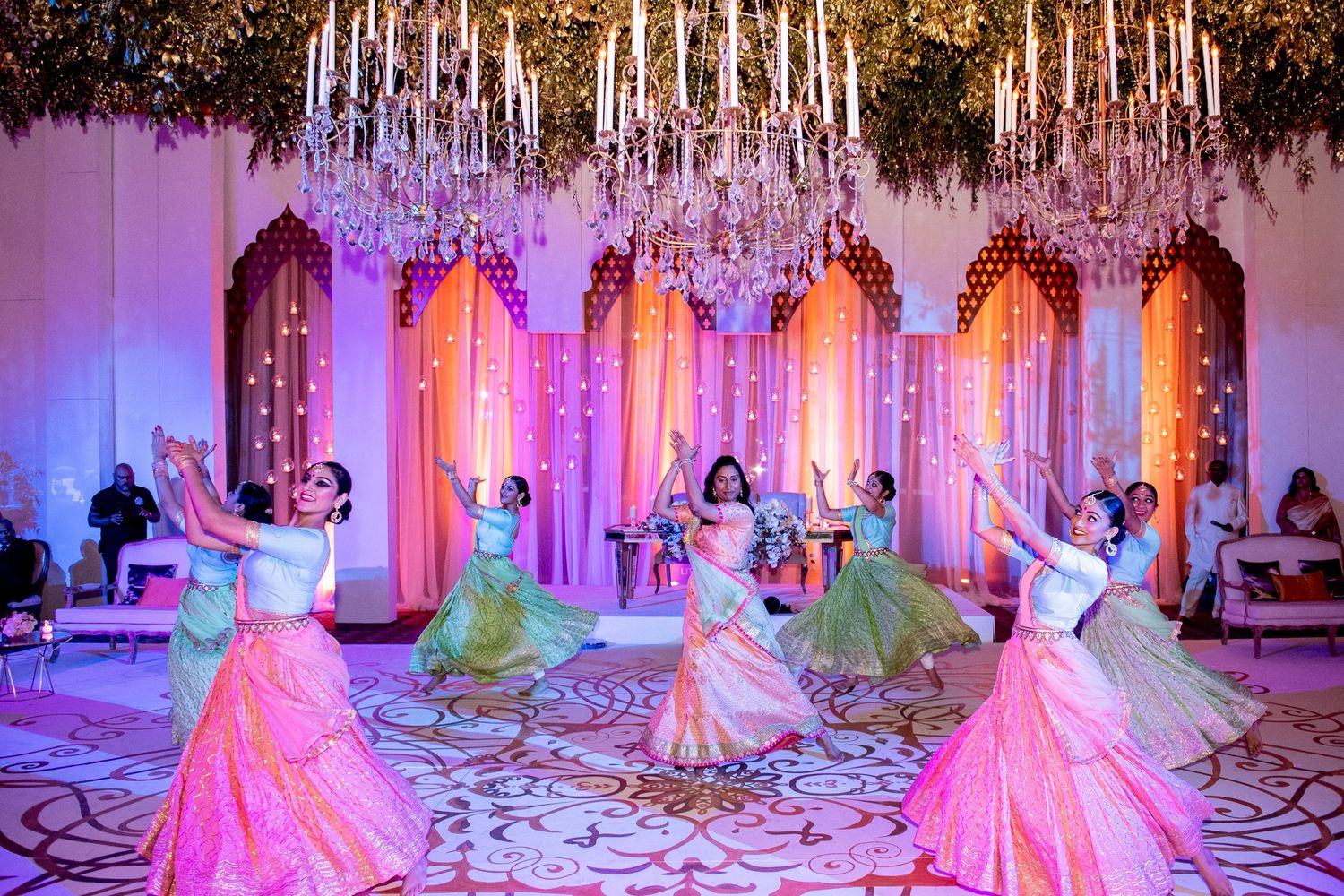 Women in colorful lehengas performing a dance at an Indian wedding celebration with crystal chandeliers.