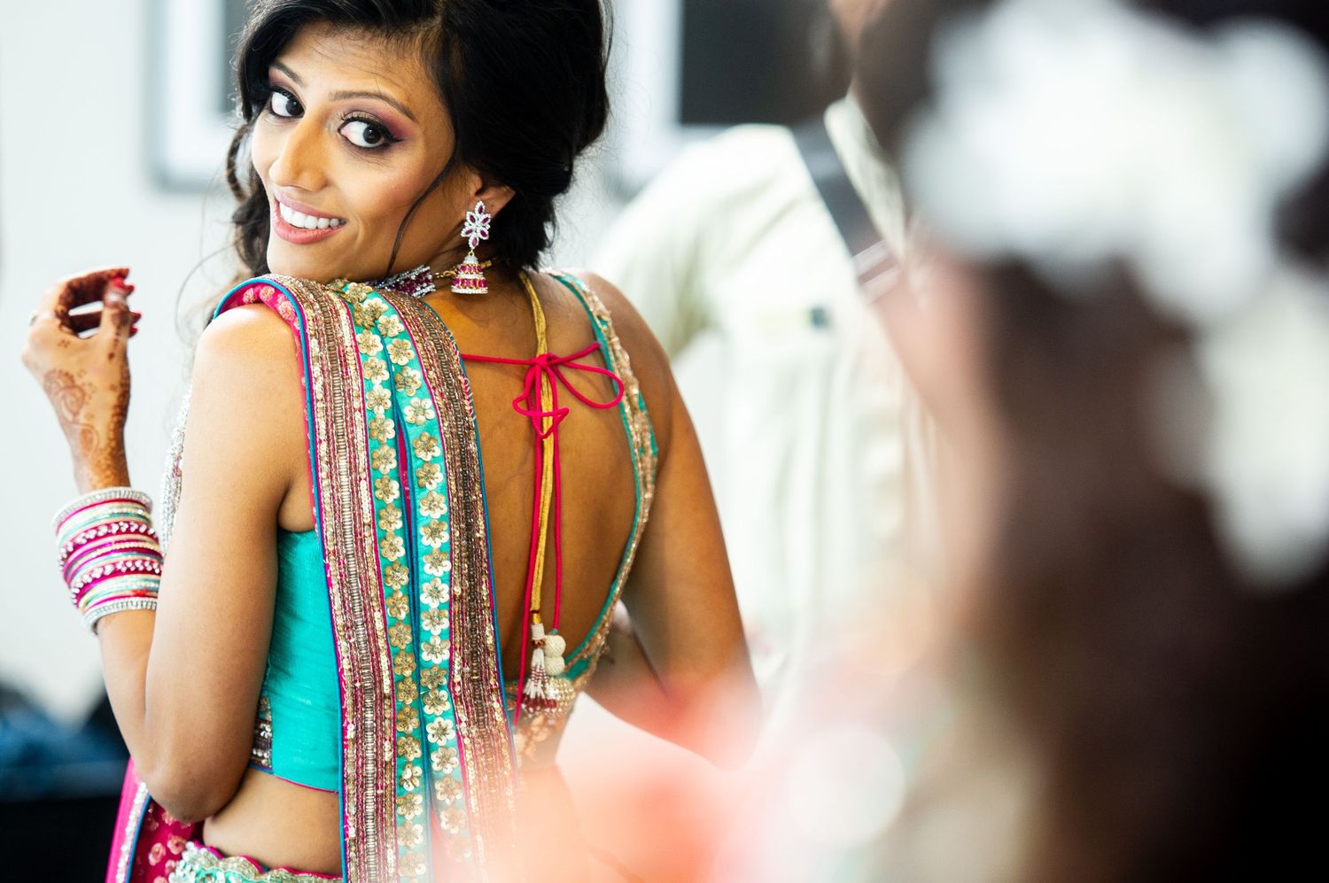 Indian bride in colorful lehenga with open back smiles over shoulder during wedding preparation.