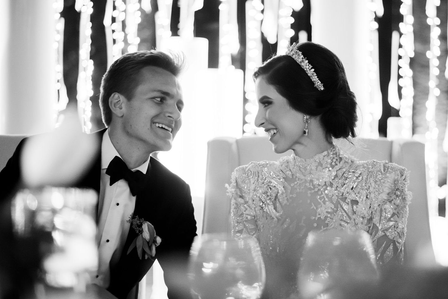 Black and white wedding photo of smiling bride in lace gown and groom in tuxedo sharing a joyful moment at reception.