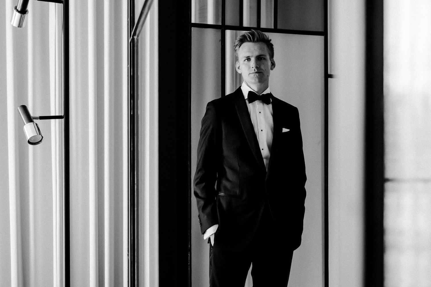 Elegant groom in black tuxedo and bow tie posing in modern interior black and white photo.