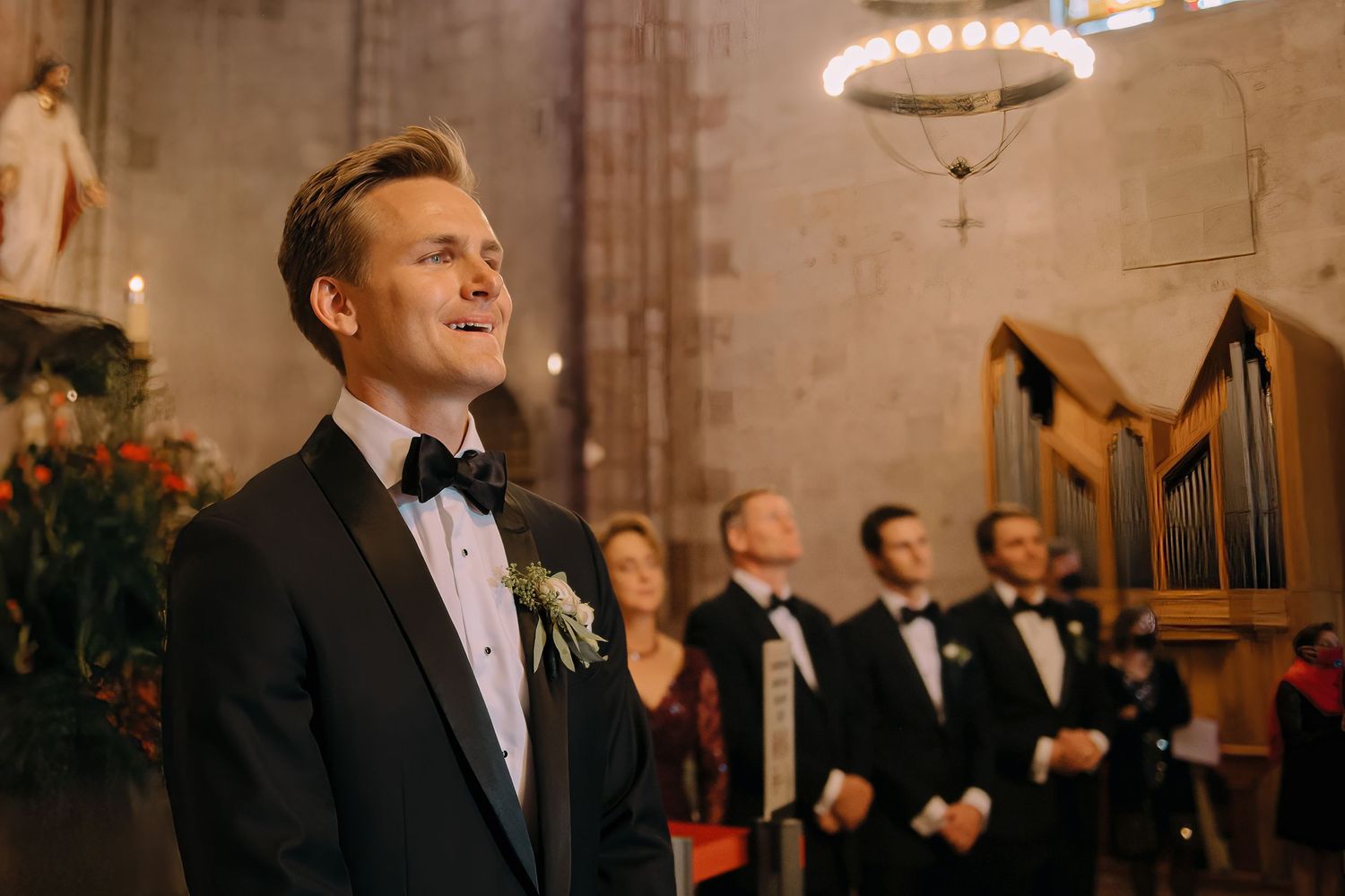 Groom in black tuxedo with boutonniere standing at church altar during wedding ceremony.